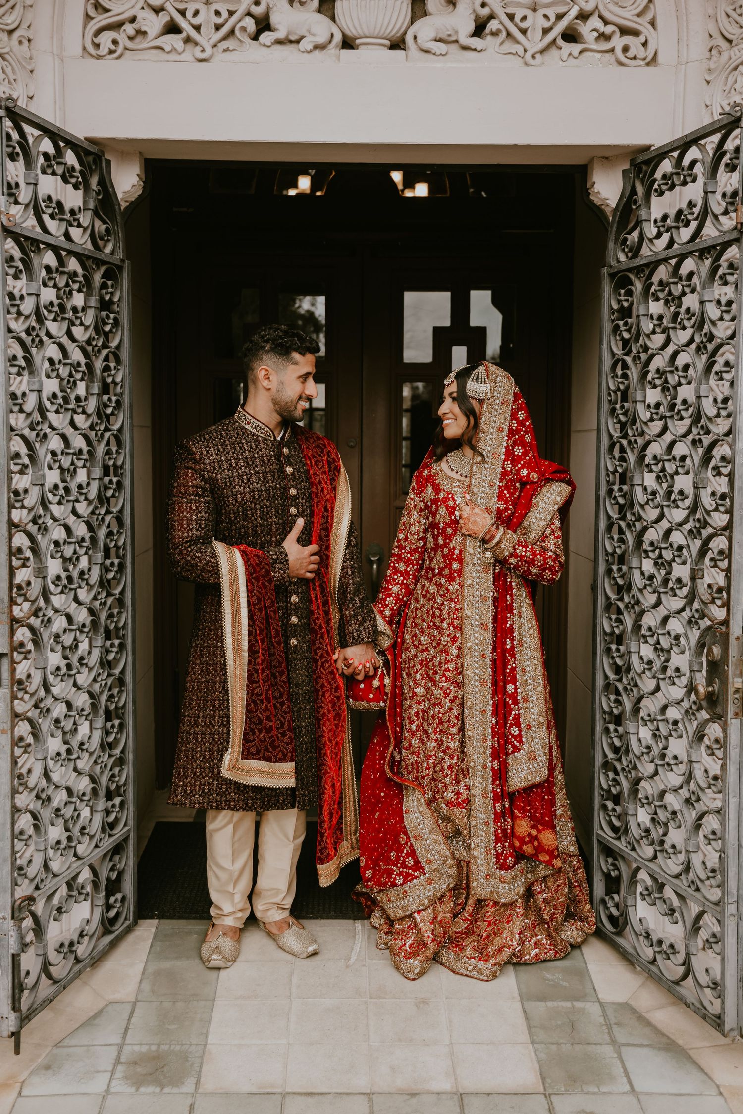 South Asian couple in traditional wedding attire posing between ornately carved door frames in elegant bridal photo.