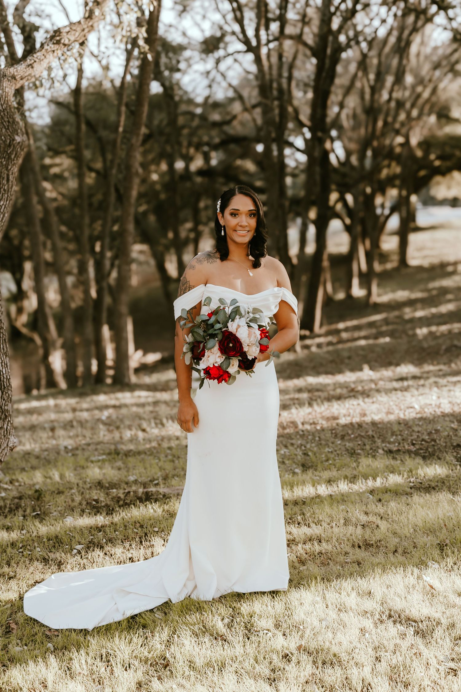 Beautiful bride in off-shoulder white wedding gown holds red and white bouquet while standing in sunlit wooded area.