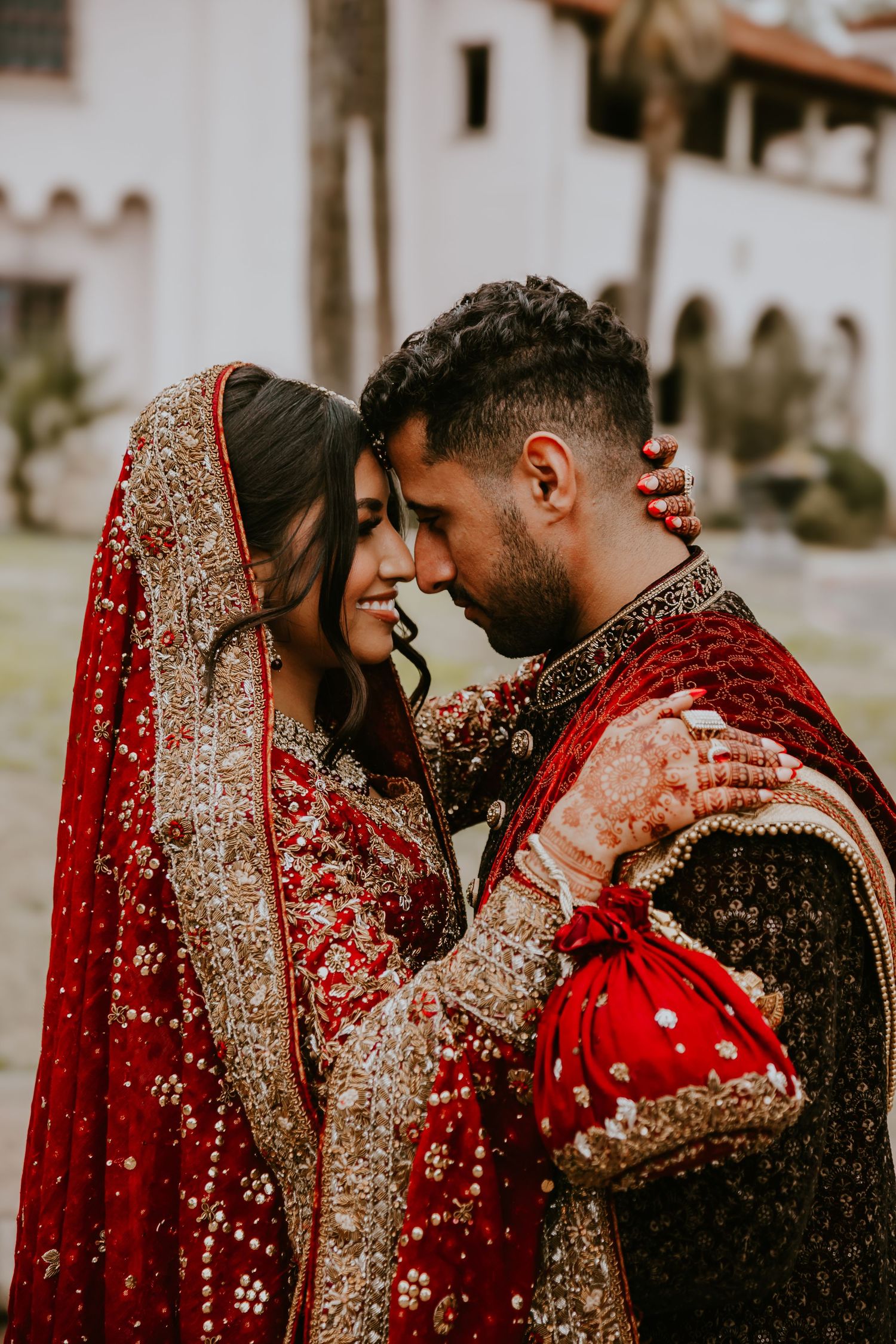 A couple in traditional South Asian wedding attire share an intimate moment in ornate red and gold outfits.