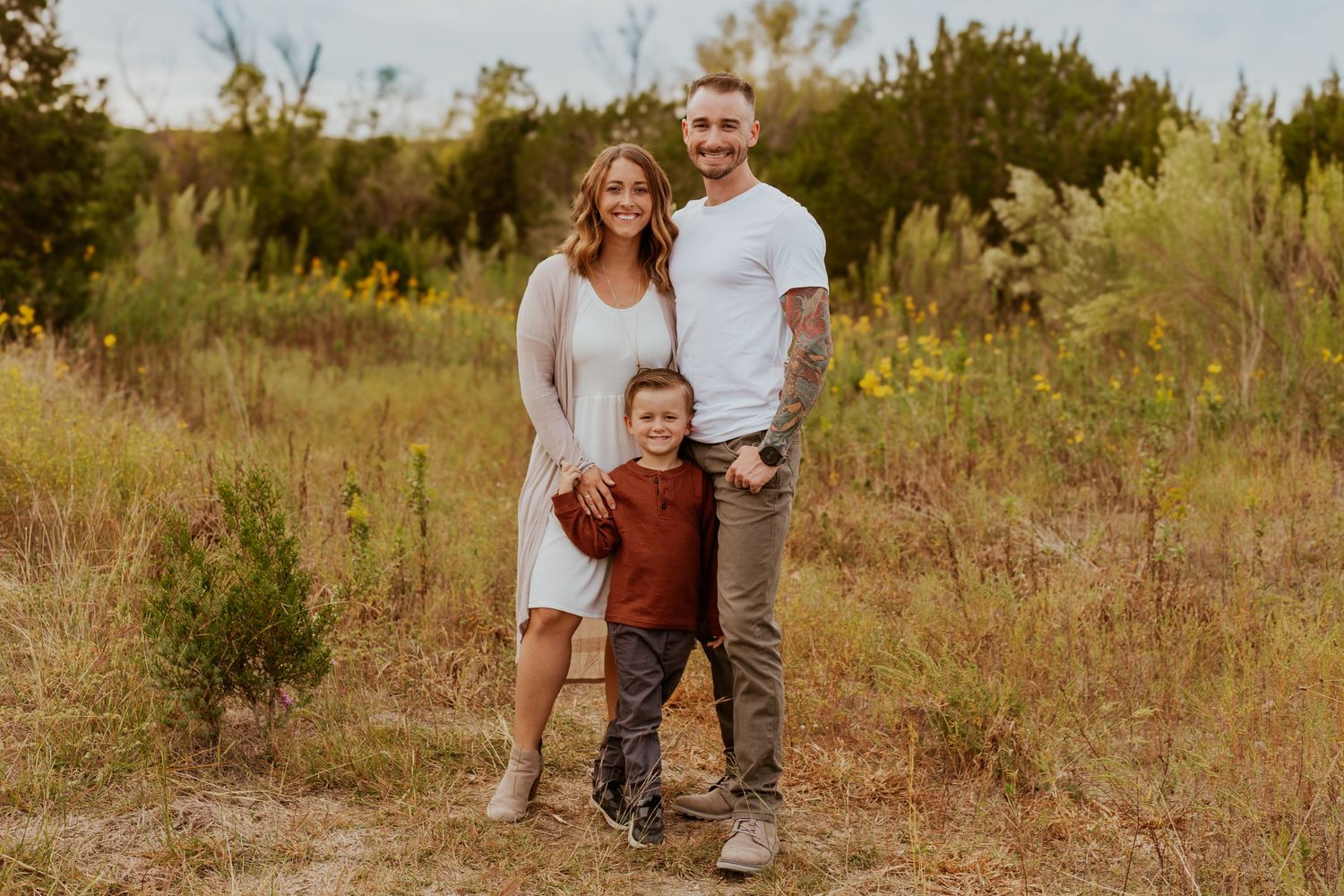 Family wearing white and brown casual outfits poses together in golden sunlit field during sunset photo session.