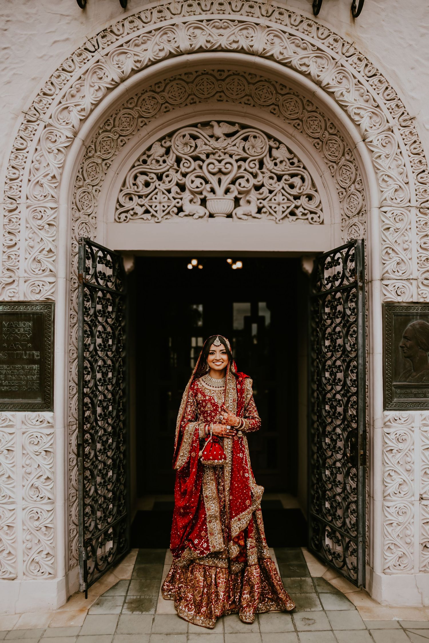 Traditional Indian bride wearing ornate red and gold lehenga standing in decorated archway with intricate stonework details.
