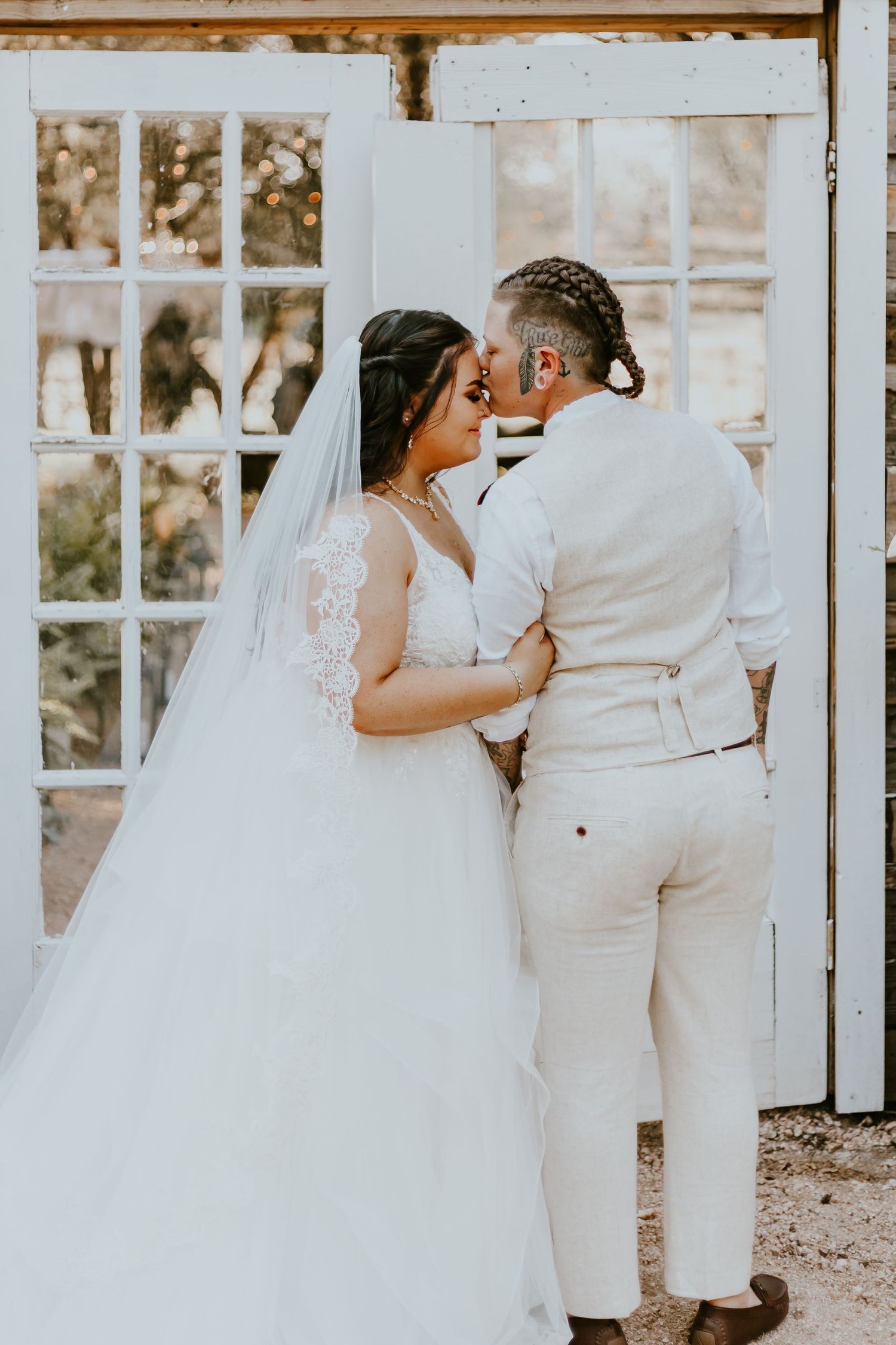 A romantic outdoor wedding portrait of a couple embracing in front of white vintage doors wearing light-colored attire.