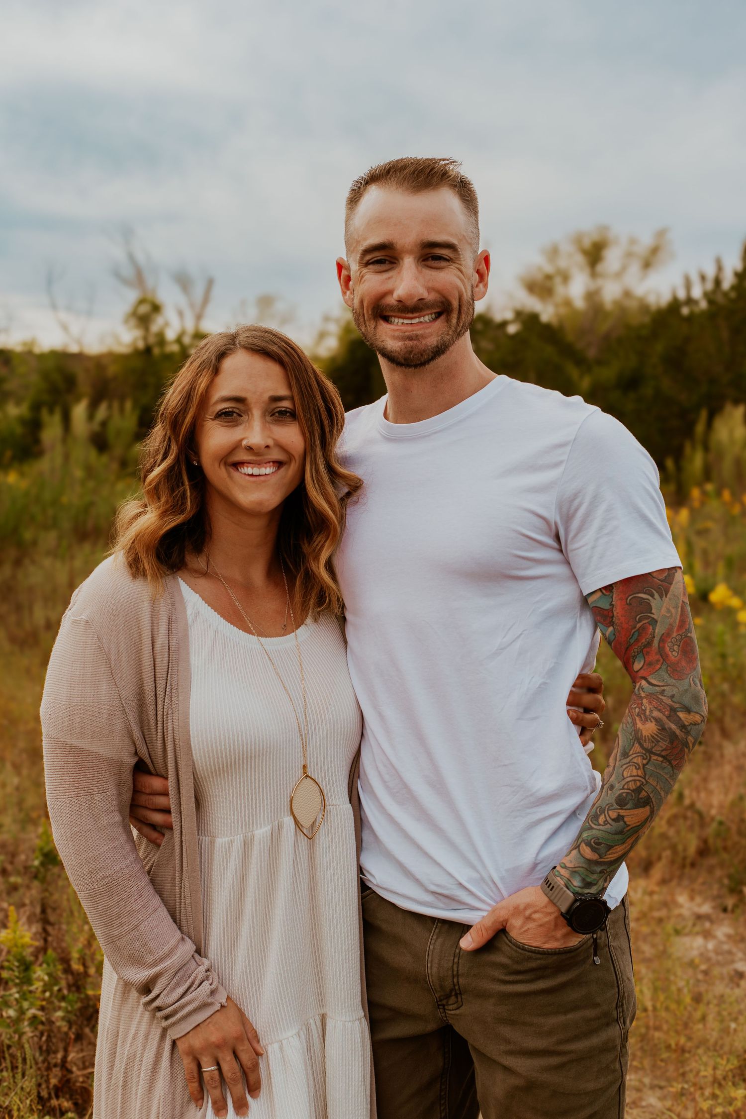 Couple poses together outdoors in a field at sunset, wearing casual clothing with natural foliage in background.