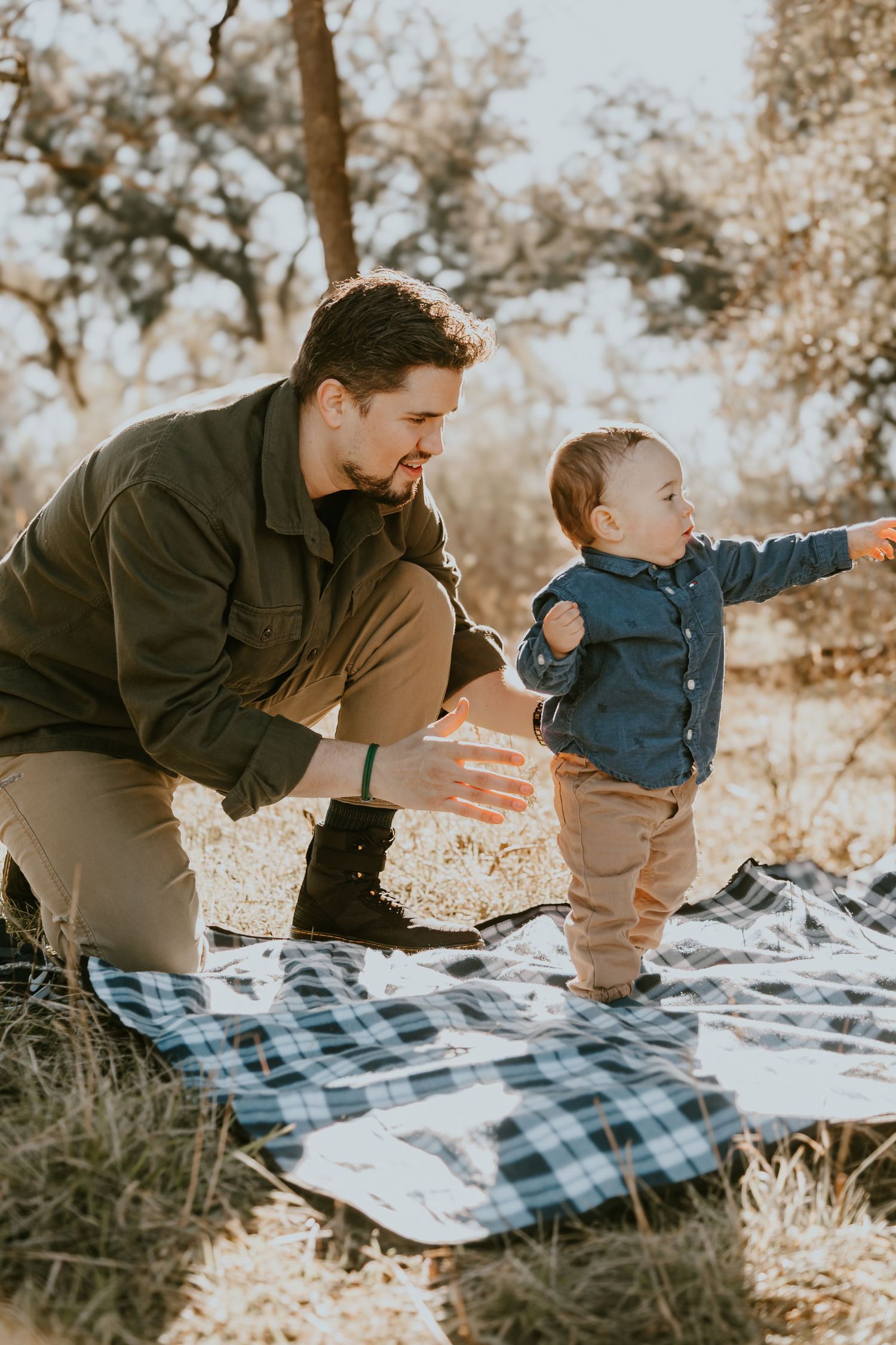 A small child points while standing on a plaid blanket outdoors during golden hour as an adult stands nearby.