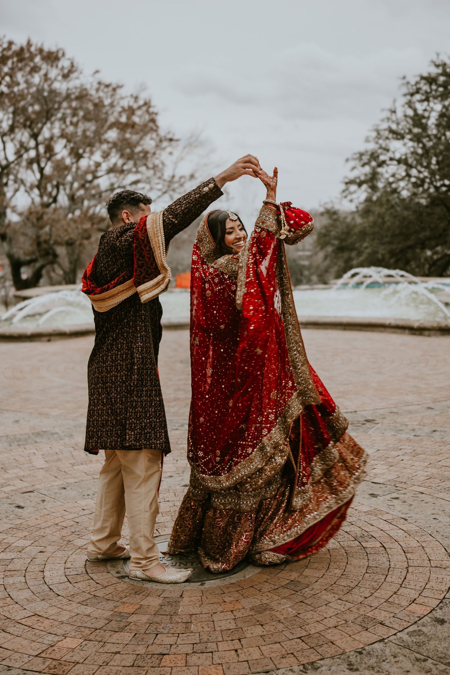 A couple in traditional South Asian wedding attire dance together on a circular brick patio surrounded by trees.