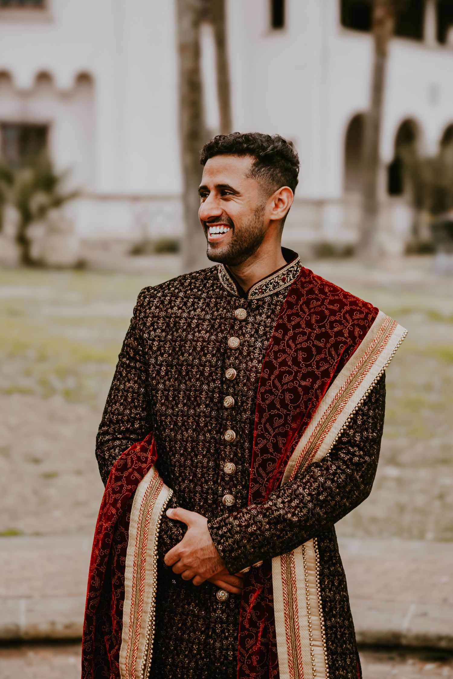 Someone wearing an ornate black and red embroidered sherwani poses during what appears to be a traditional Indian wedding.