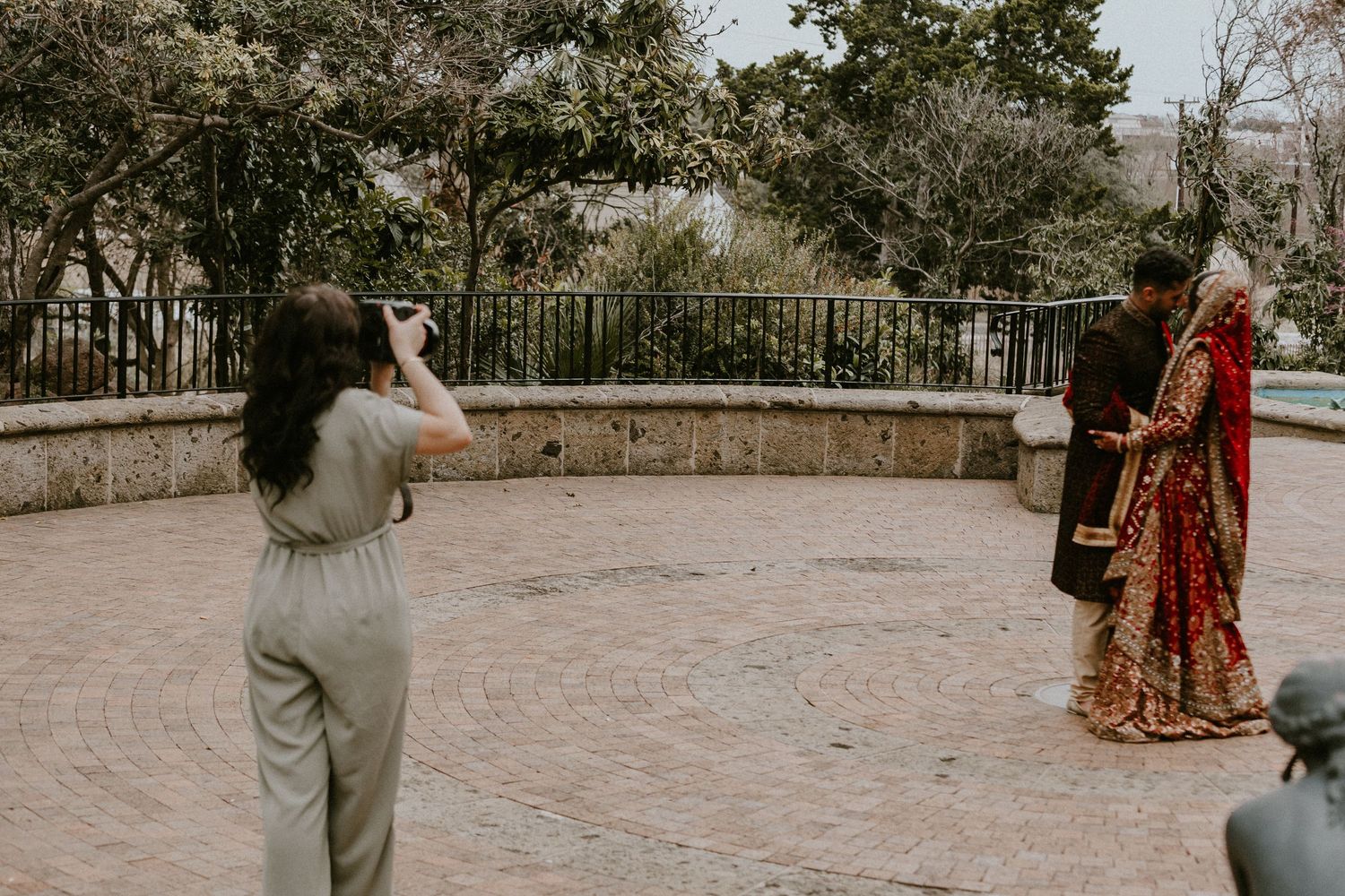 A photographer captures someone in traditional red and gold wedding attire in an outdoor courtyard setting.