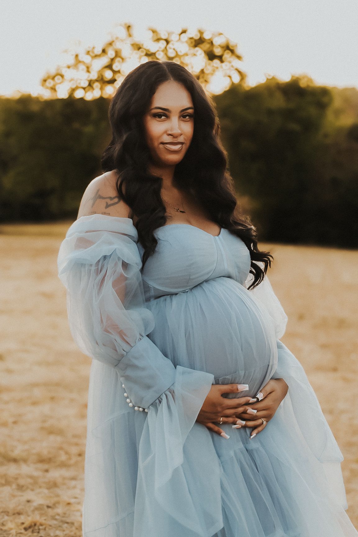 A person in a light blue tulle maternity gown poses in a sunlit field during golden hour.