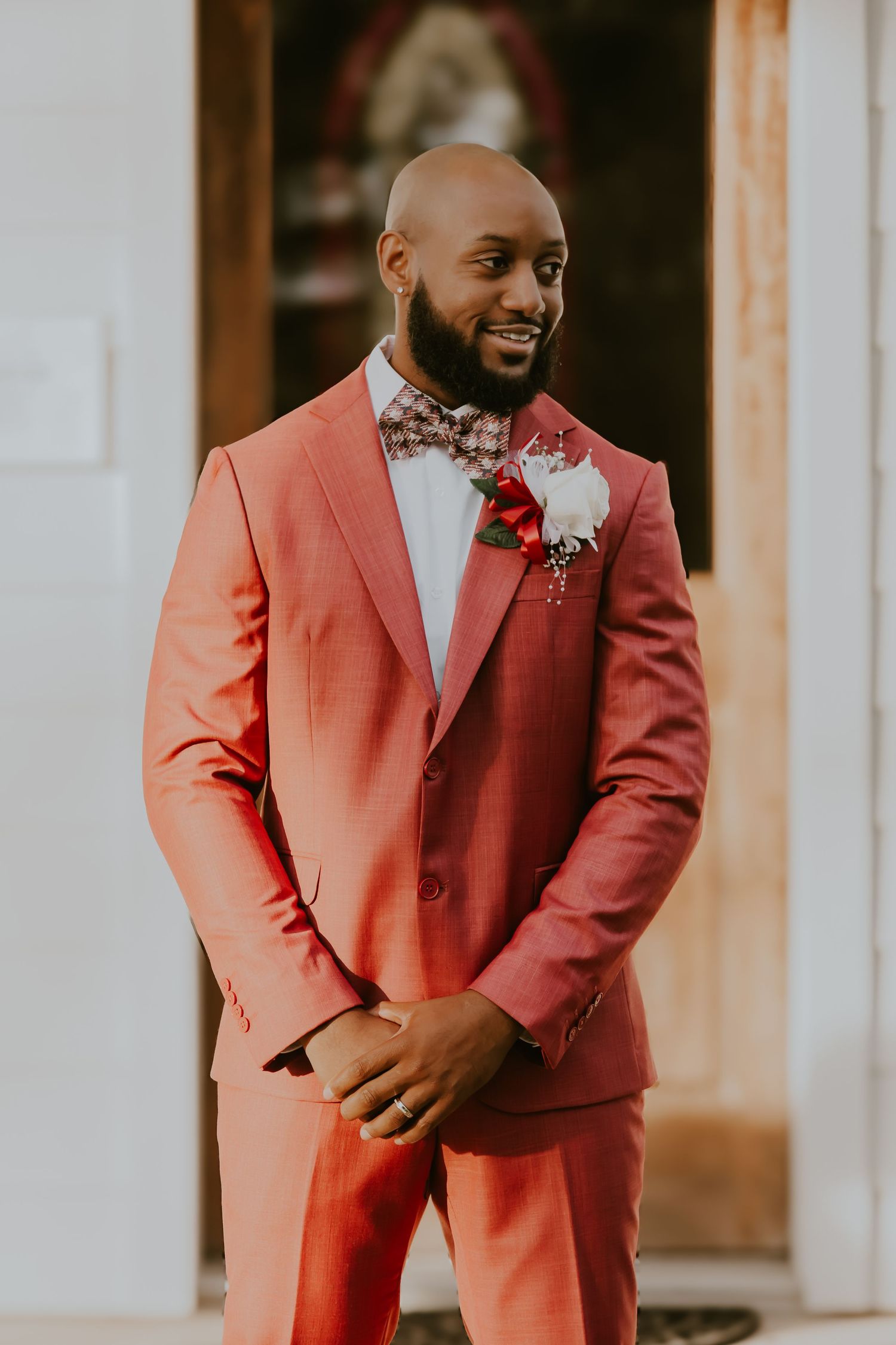 A stylish groom wearing a salmon pink suit with bowtie and white boutonniere smiles warmly at a wedding.