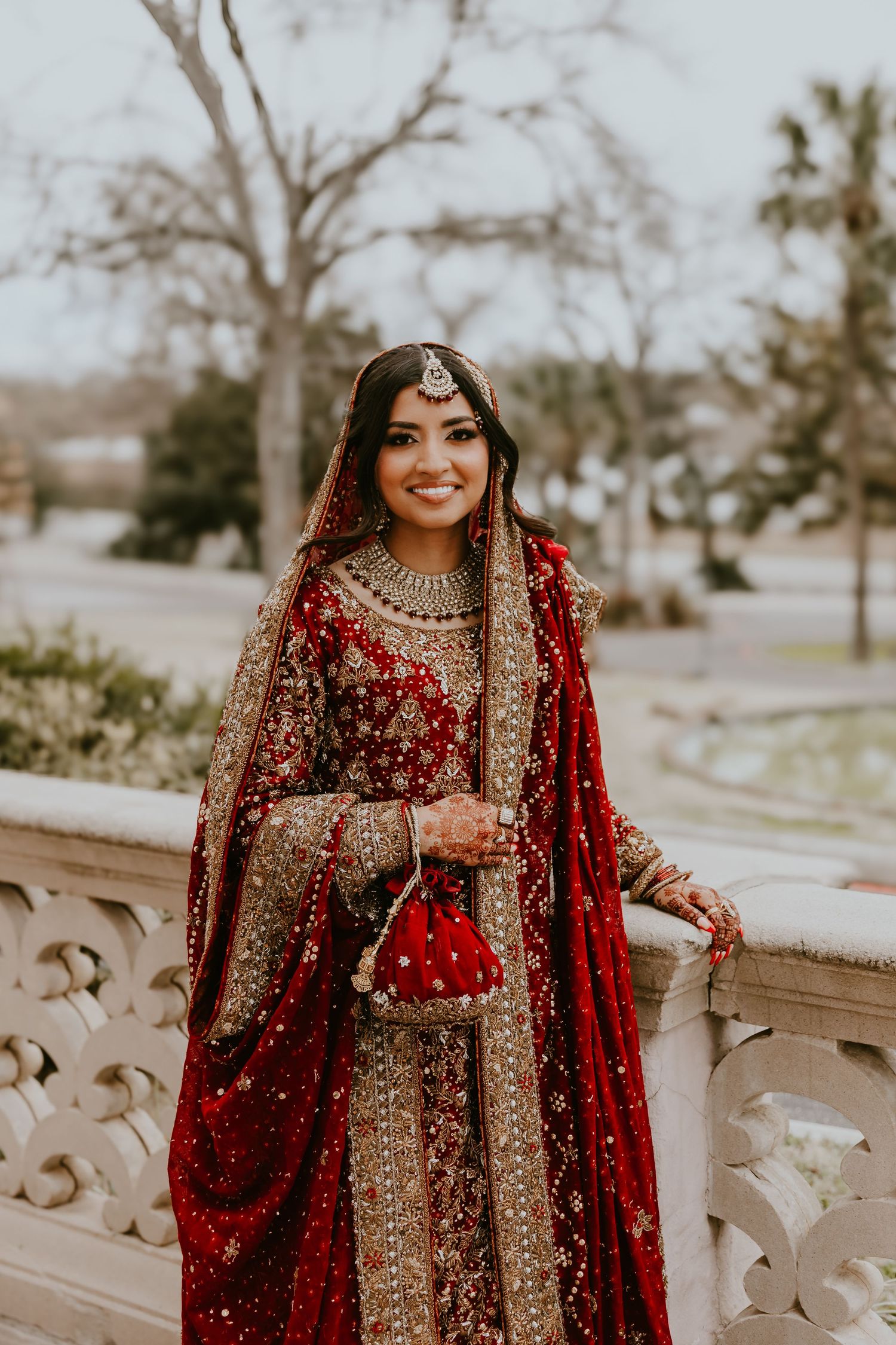 Traditional South Asian bride wearing an ornate red and gold lehenga with detailed embroidery and jewelry outdoors.