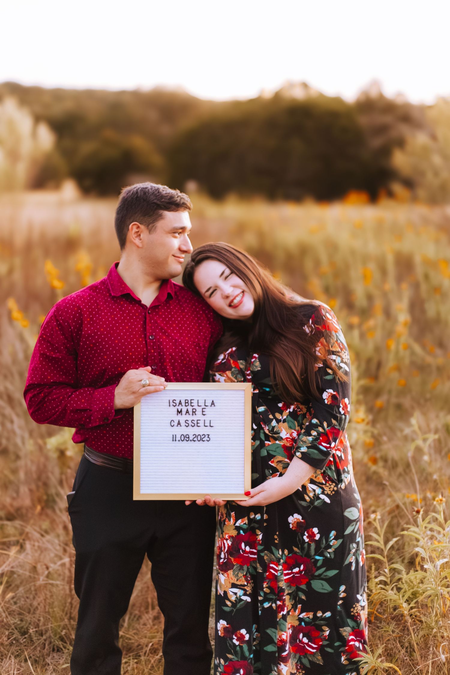 A couple poses in a golden field at sunset holding a letterboard sign during an engagement photo session.