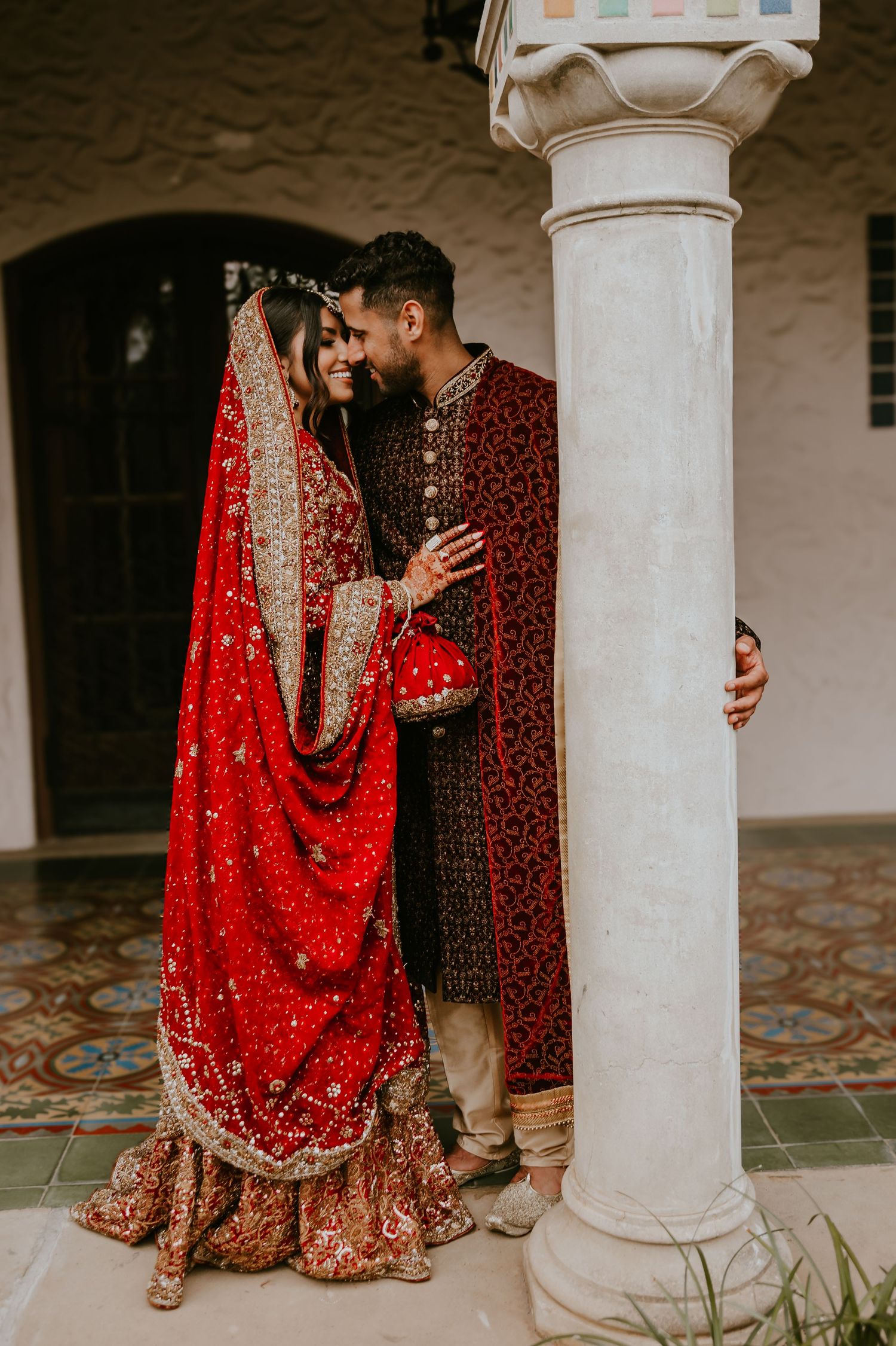 Traditional South Asian wedding couple in ornate red and maroon attire embrace by white pillar in romantic moment.