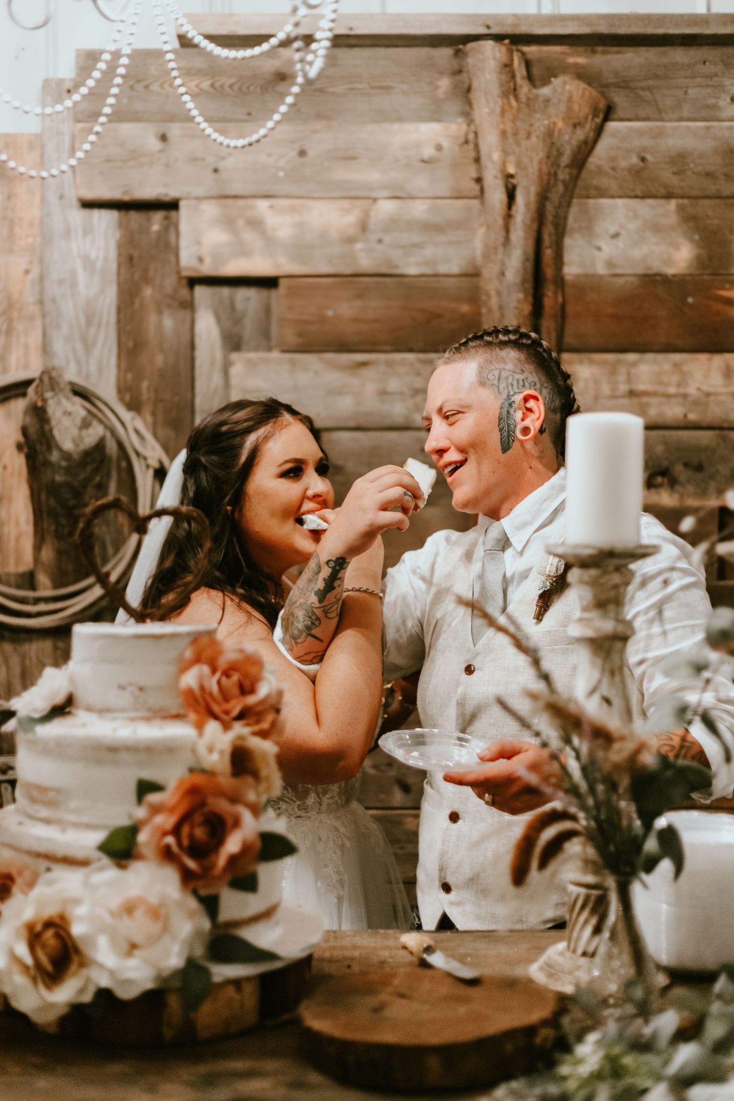 Couple shares joyful moment while cutting wedding cake at rustic reception with roses and string lights.