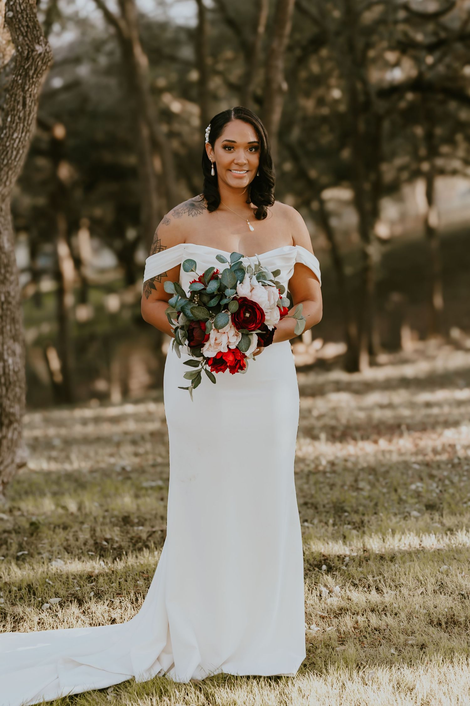 A bride poses outdoors in a fitted white wedding dress holding a romantic burgundy and white floral bouquet.