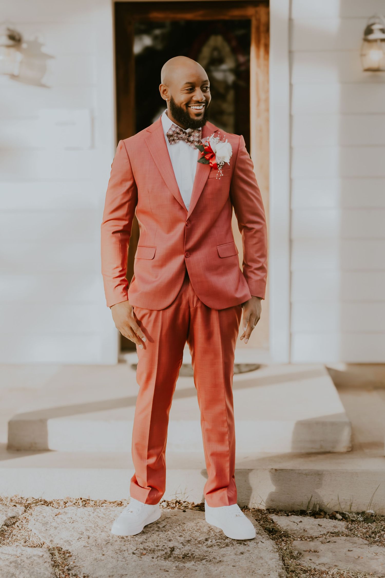Well-dressed groom in a salmon pink suit with bow tie and boutonniere stands outdoors against white walls.