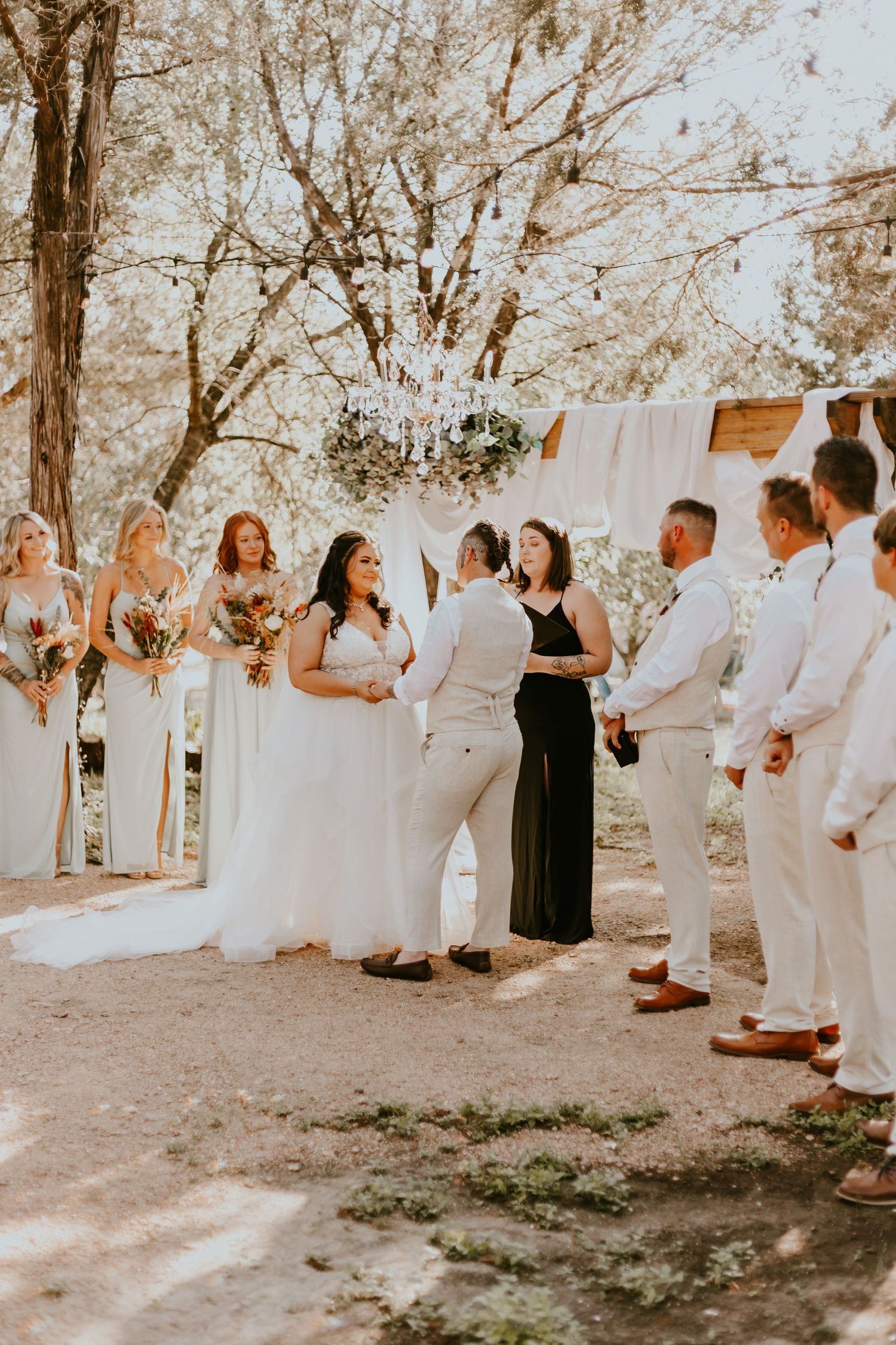 A warm-toned outdoor wedding ceremony with bridesmaids in blush dresses and groomsmen in white under a floral chandelier.
