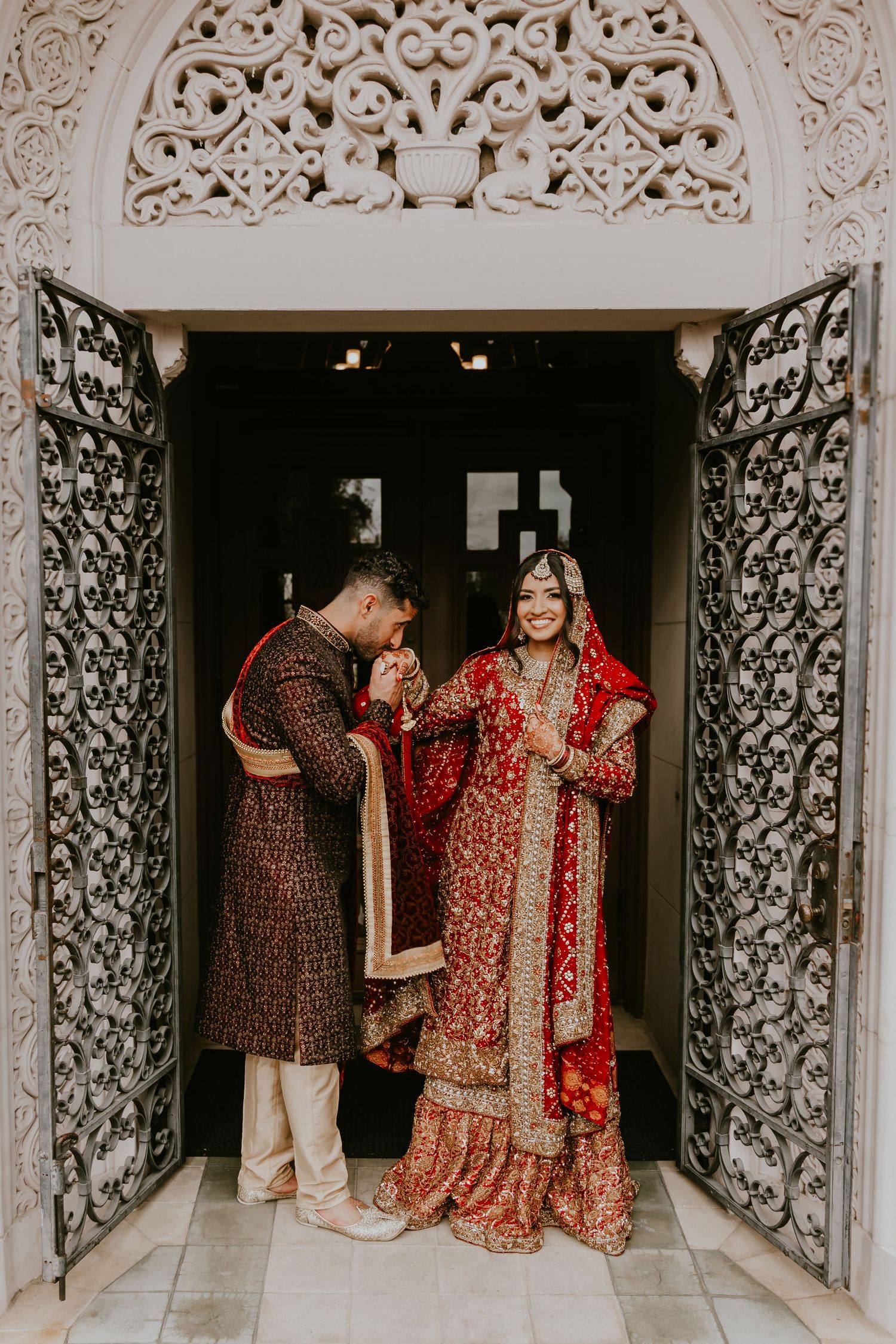 Traditional wedding couple in ornate red and brown attire stand in doorway with decorative carved stone archway and metal gates.