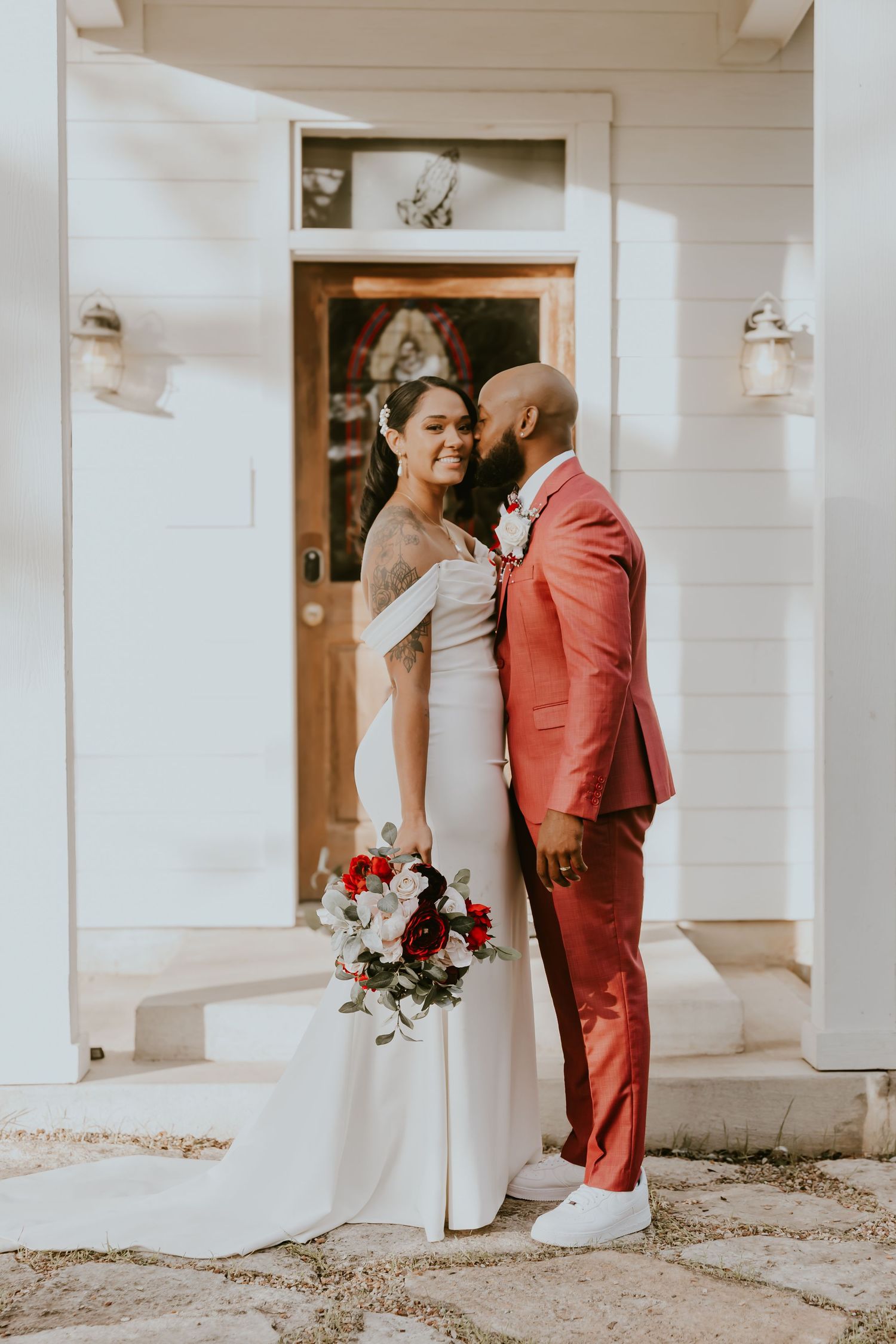 Newlywed couple embraces outside white church entrance, bride in white dress holding red bouquet, groom in coral suit.