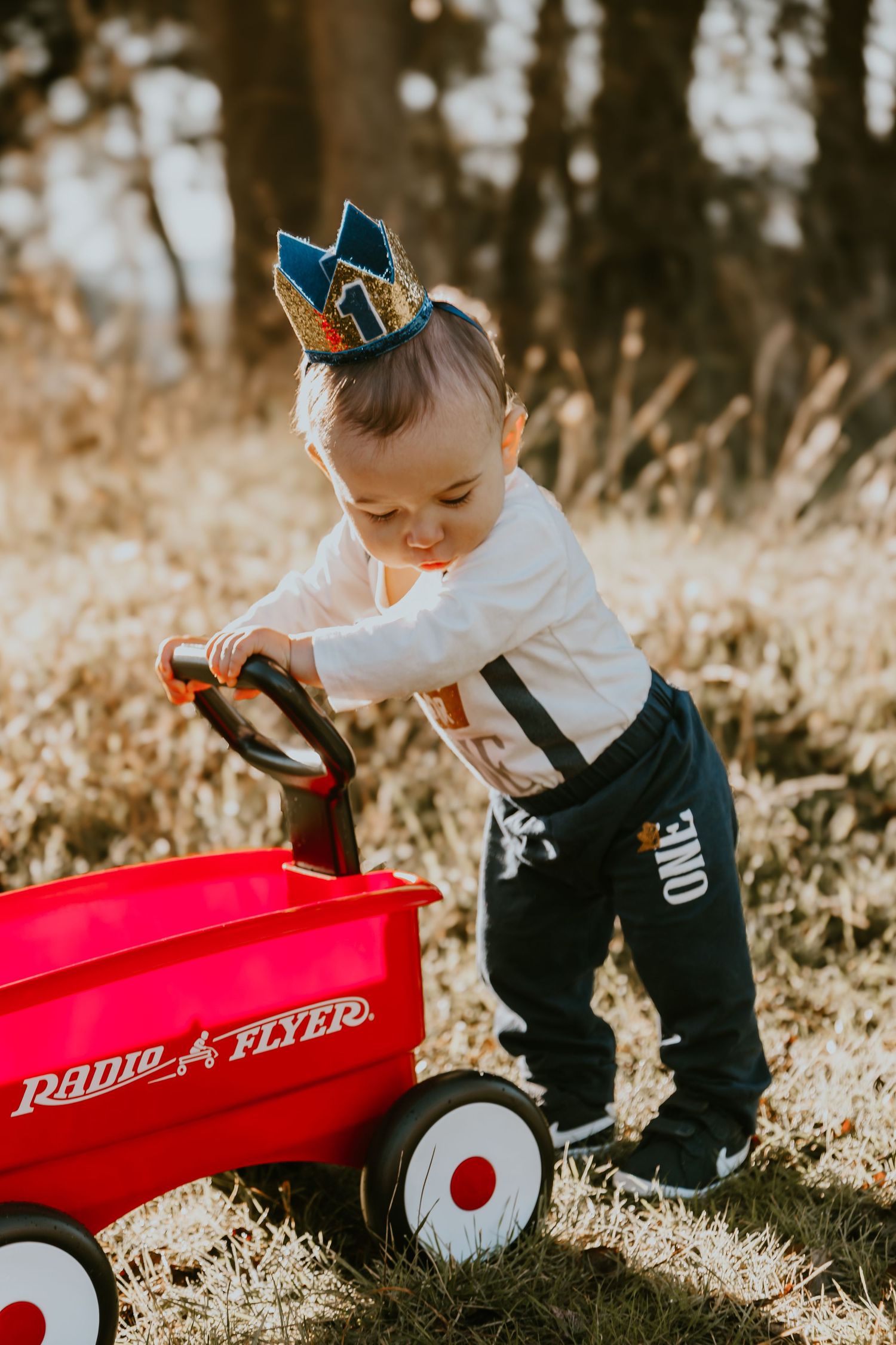 Baby wearing blue crown plays with classic red Radio Flyer wagon in autumn sunlit outdoor setting.