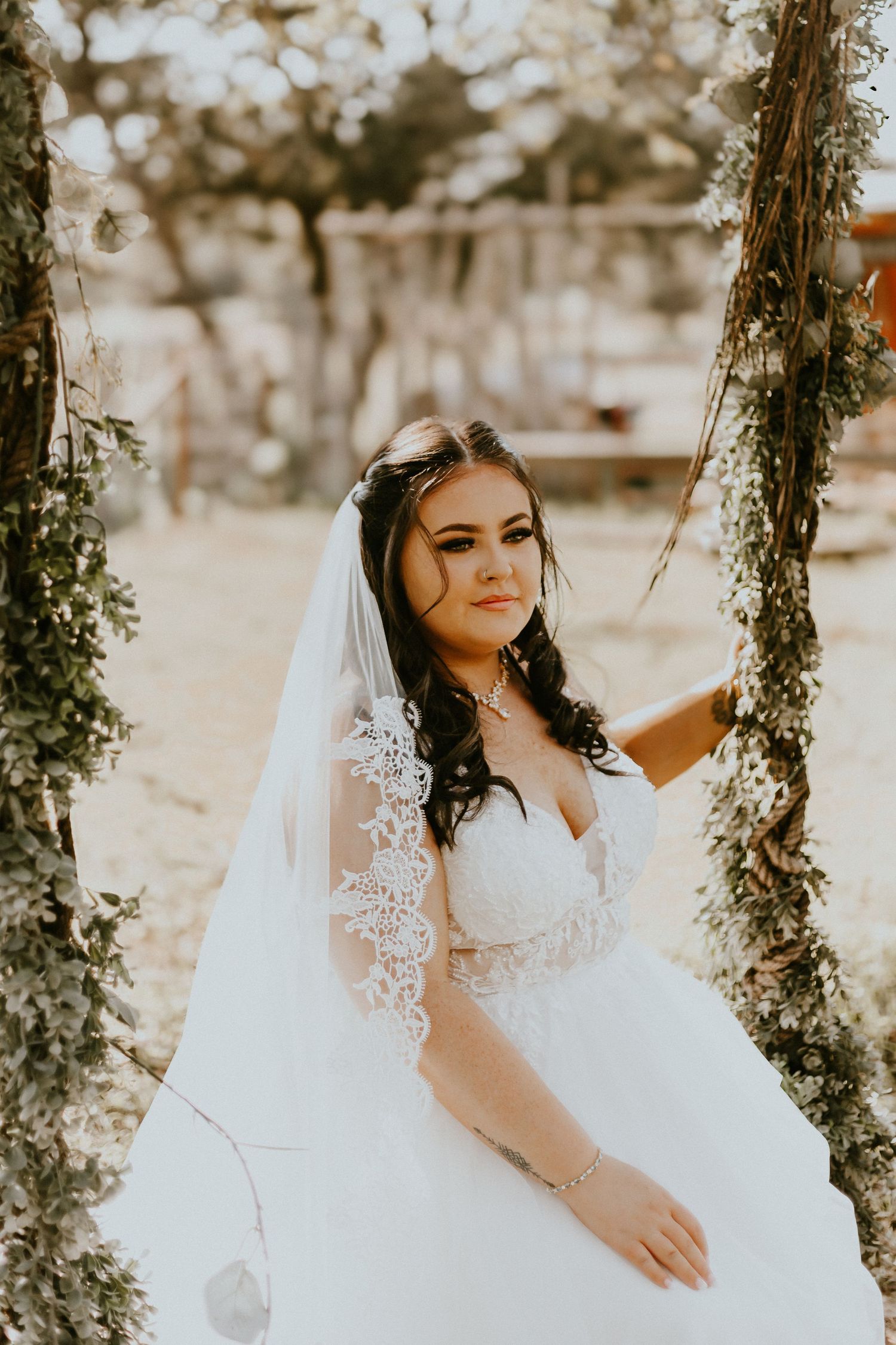 Bride in white lace wedding dress sits on garden swing decorated with greenery in warm sunlight.