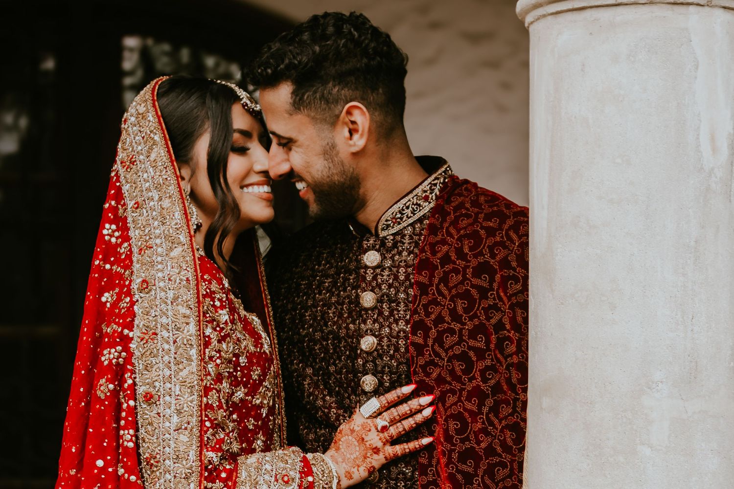 A couple in traditional South Asian wedding attire shares an intimate moment against a white pillar.