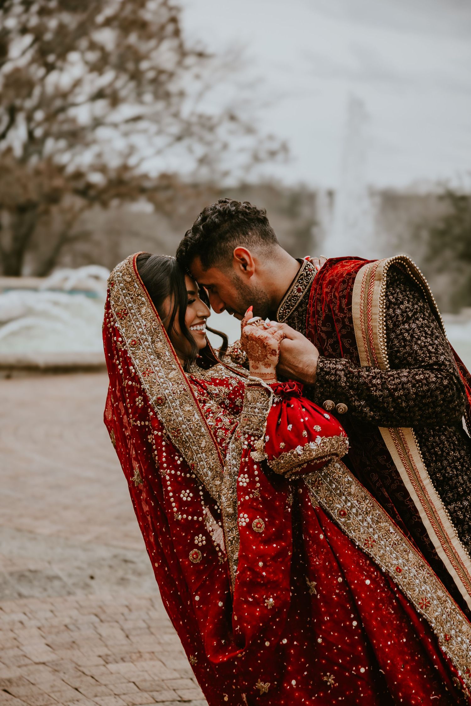 A romantic couple in traditional South Asian wedding attire embrace while wearing ornate red and gold lehenga and sherwani.