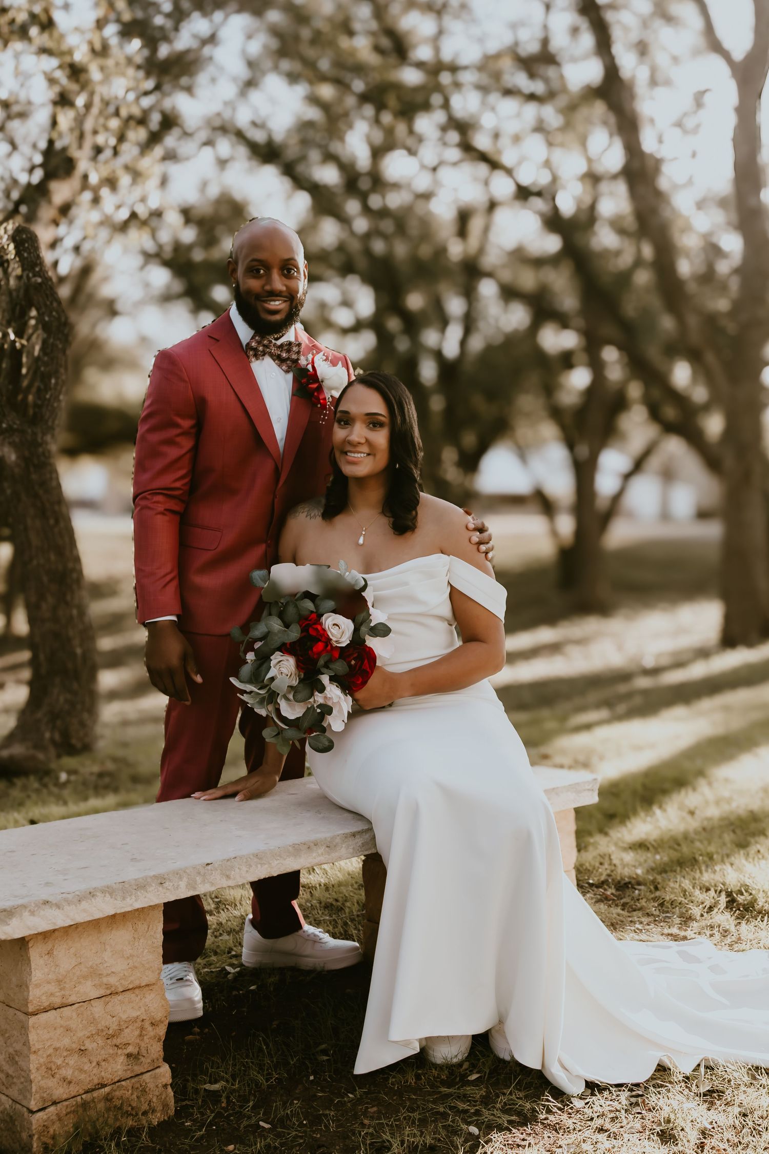 A beautiful outdoor wedding photo with a couple sitting on a stone bench in warm sunlight among trees.
