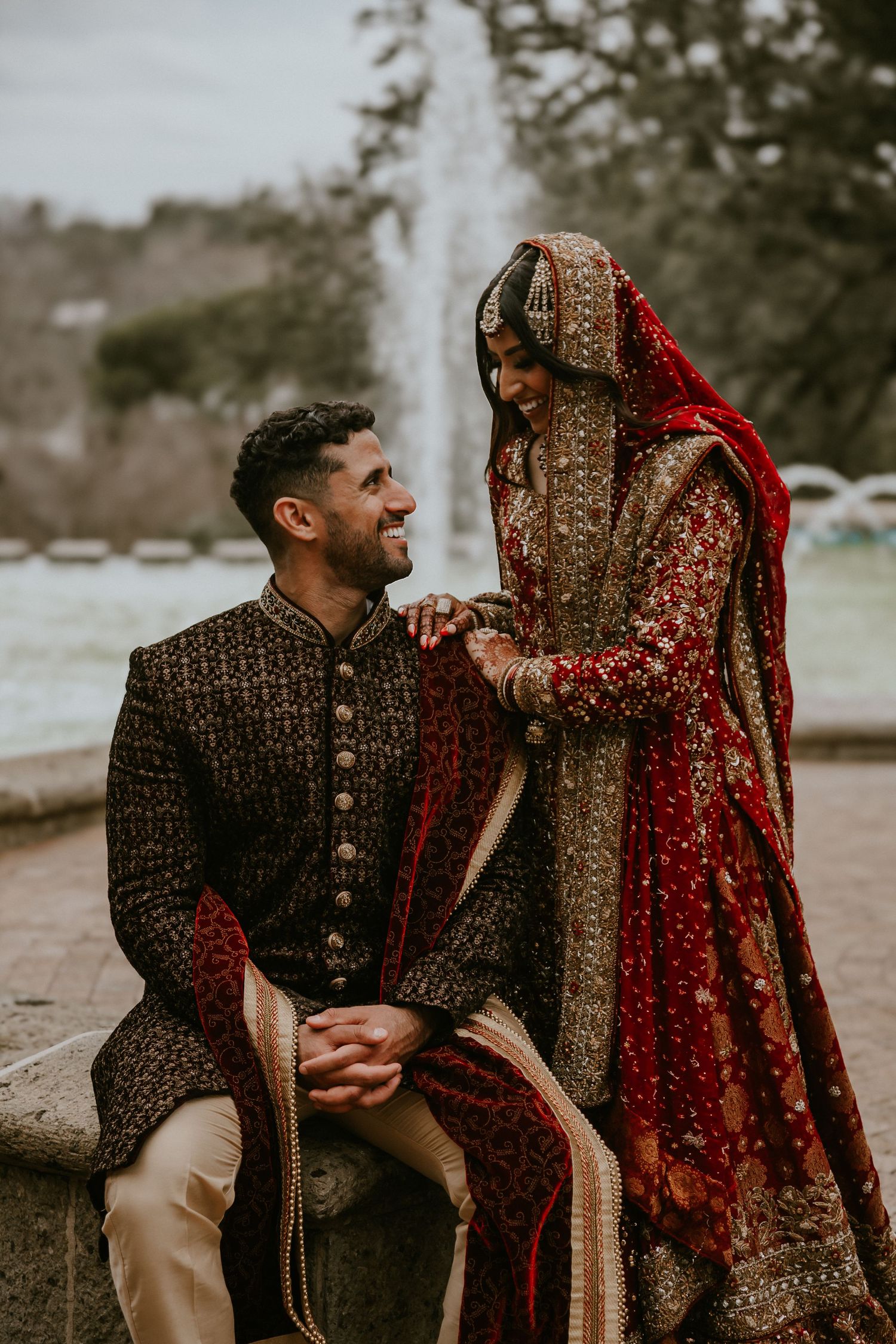 A couple in traditional South Asian wedding attire shares an intimate moment outdoors with ornate embroidered details.