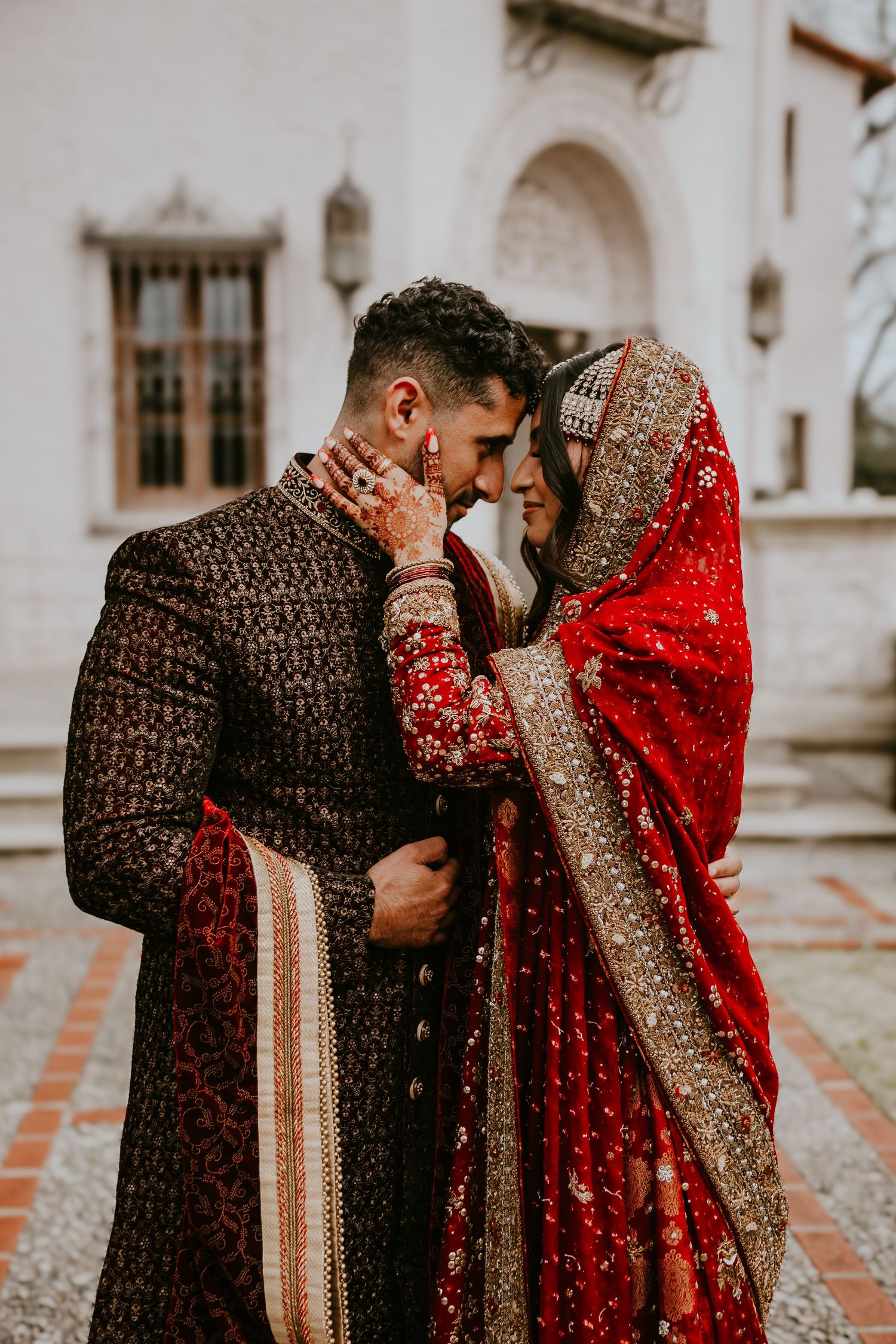 A couple in traditional South Asian wedding attire share an intimate moment in ornate red and black ethnic dress.