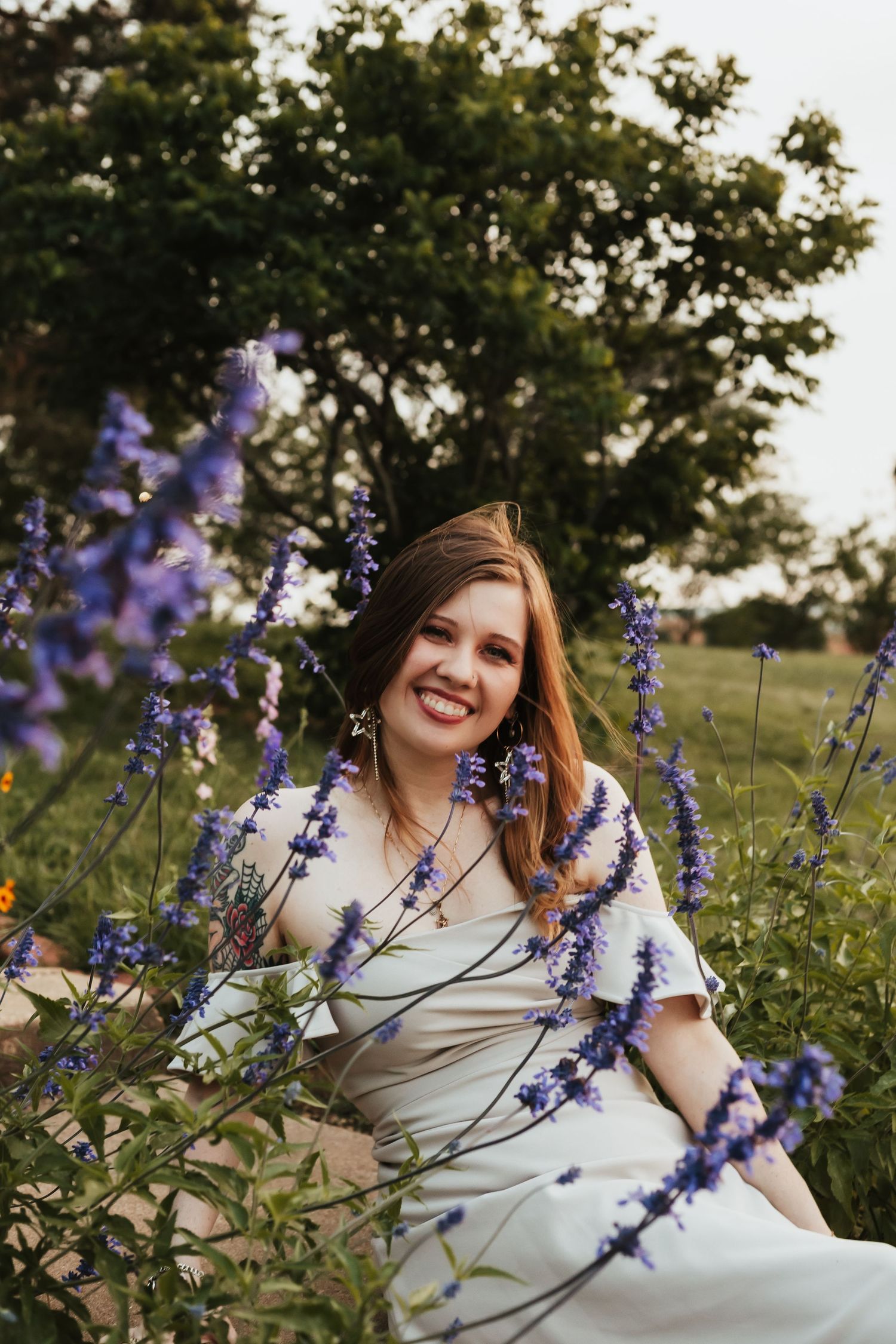 Person in white dress smiles while surrounded by blooming purple flowers in garden setting.