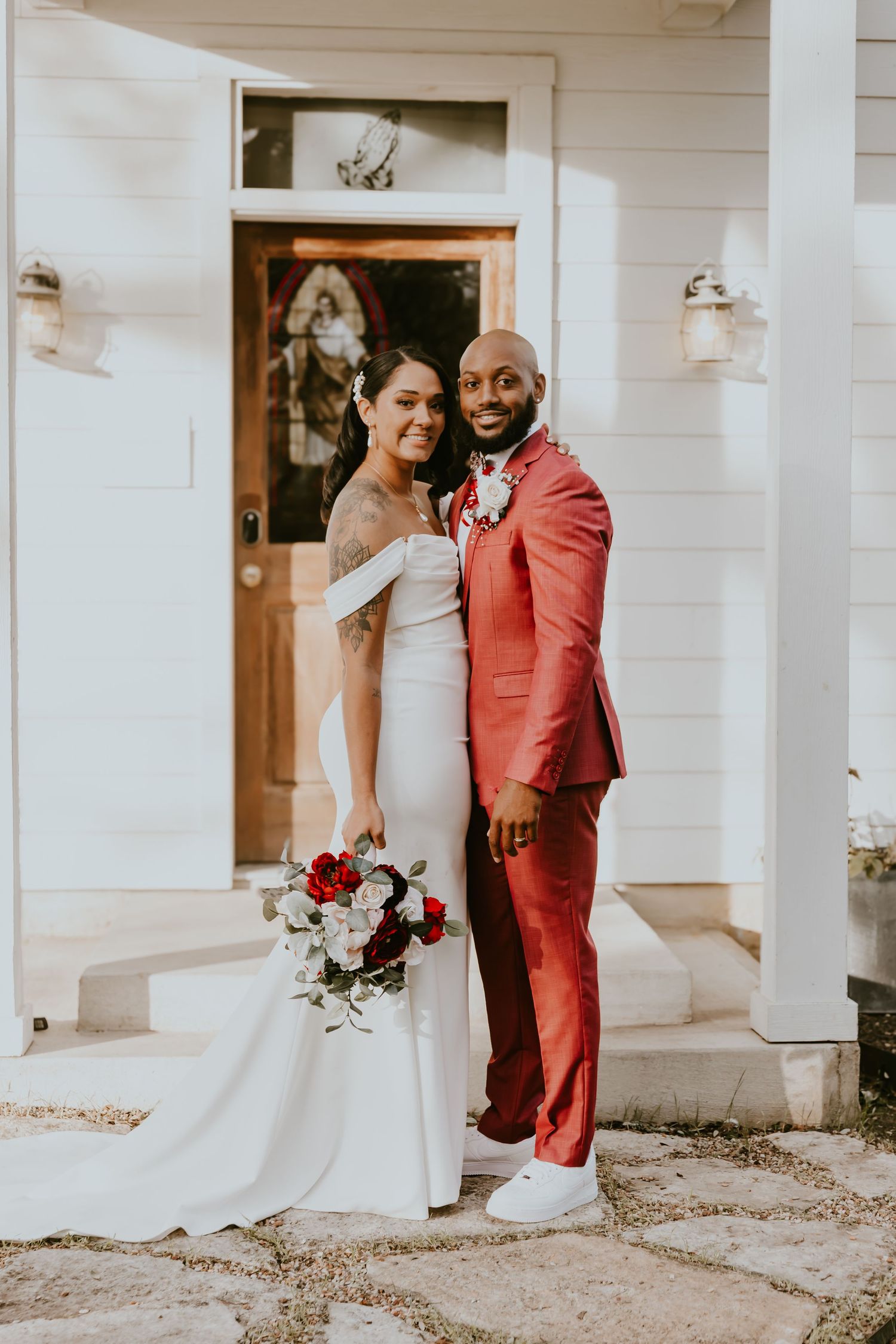 Elegant couple poses in wedding attire, with bride in white dress holding bouquet and groom in striking coral suit.