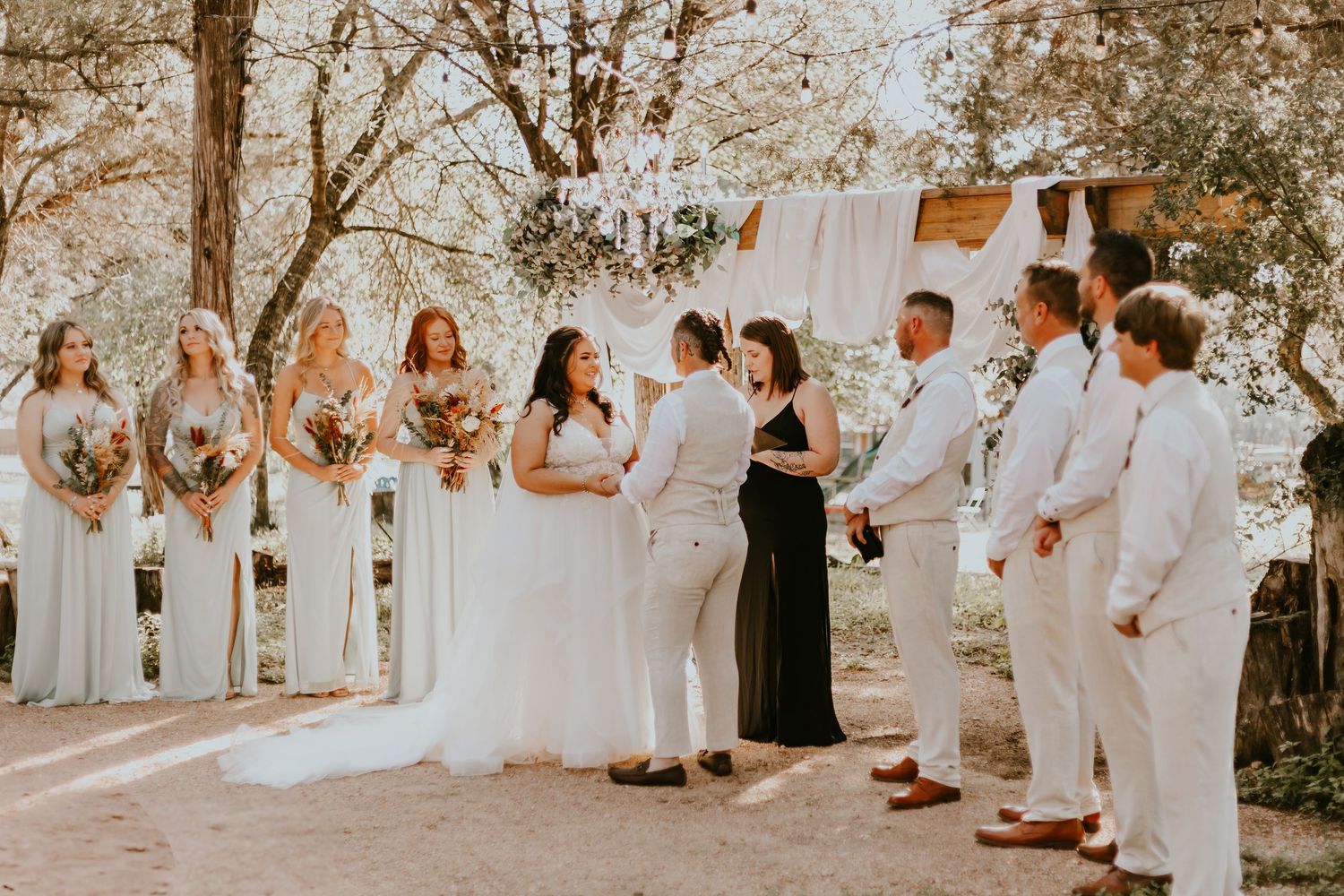Wedding party stands in sunlit outdoor ceremony with bridesmaids in gray and groomsmen in white suits.