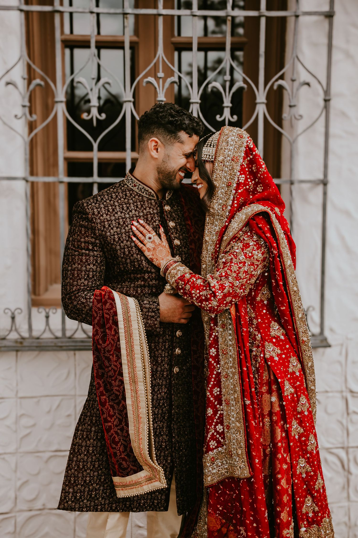 Traditional South Asian wedding couple in ornate attire share an intimate moment, with bride in red lehenga and groom in brown sherwani.