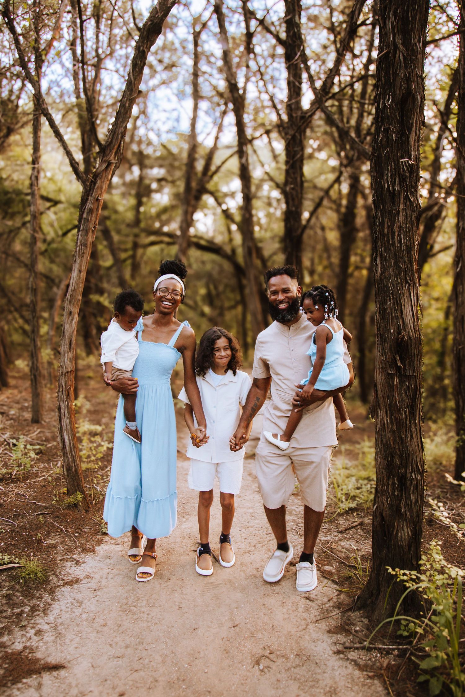 A family walks together on a dirt trail through a wooded forest area on a sunny day.