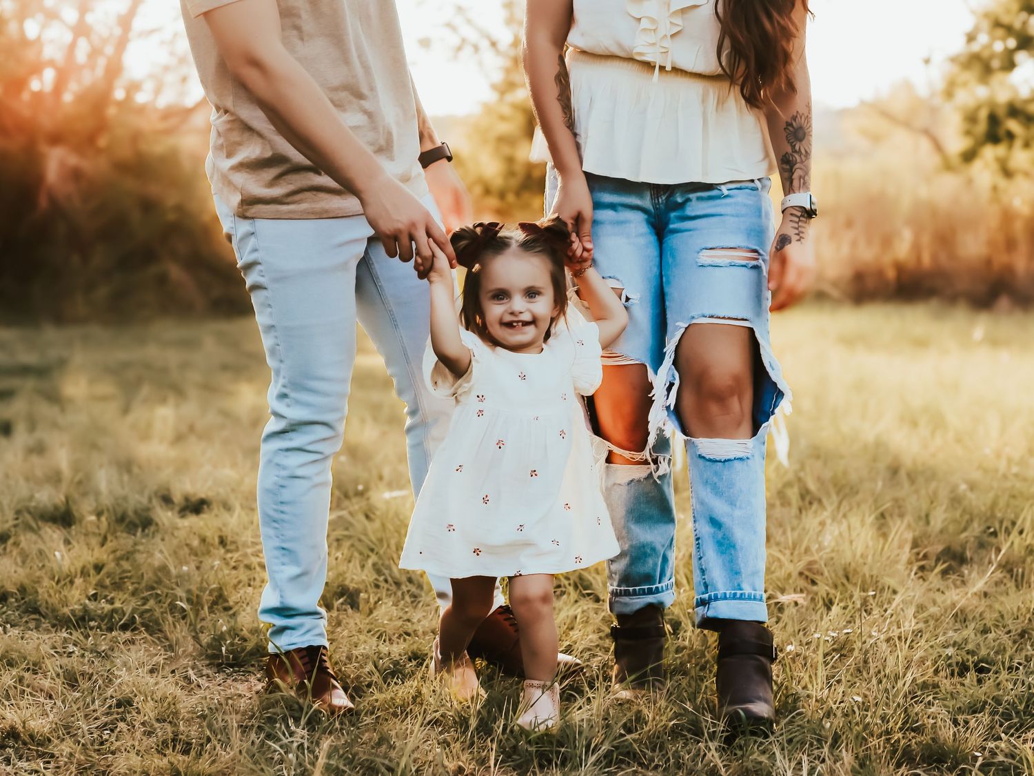 A child walks barefoot in a sundress holding hands with adults wearing jeans in a sunlit grassy field at sunset.