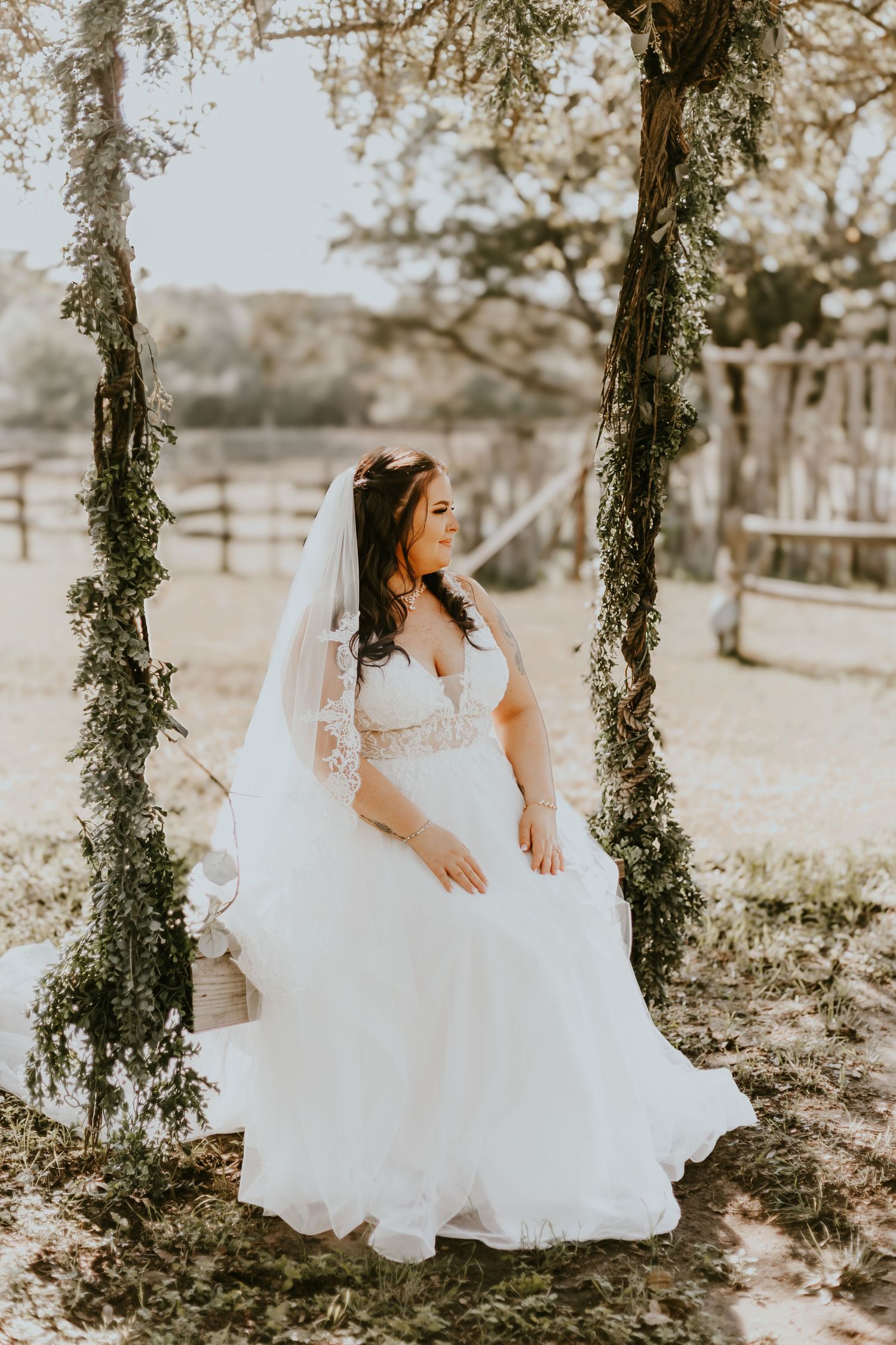 A bride in a white lace wedding dress and veil stands between two vine-covered posts in a rustic outdoor setting.