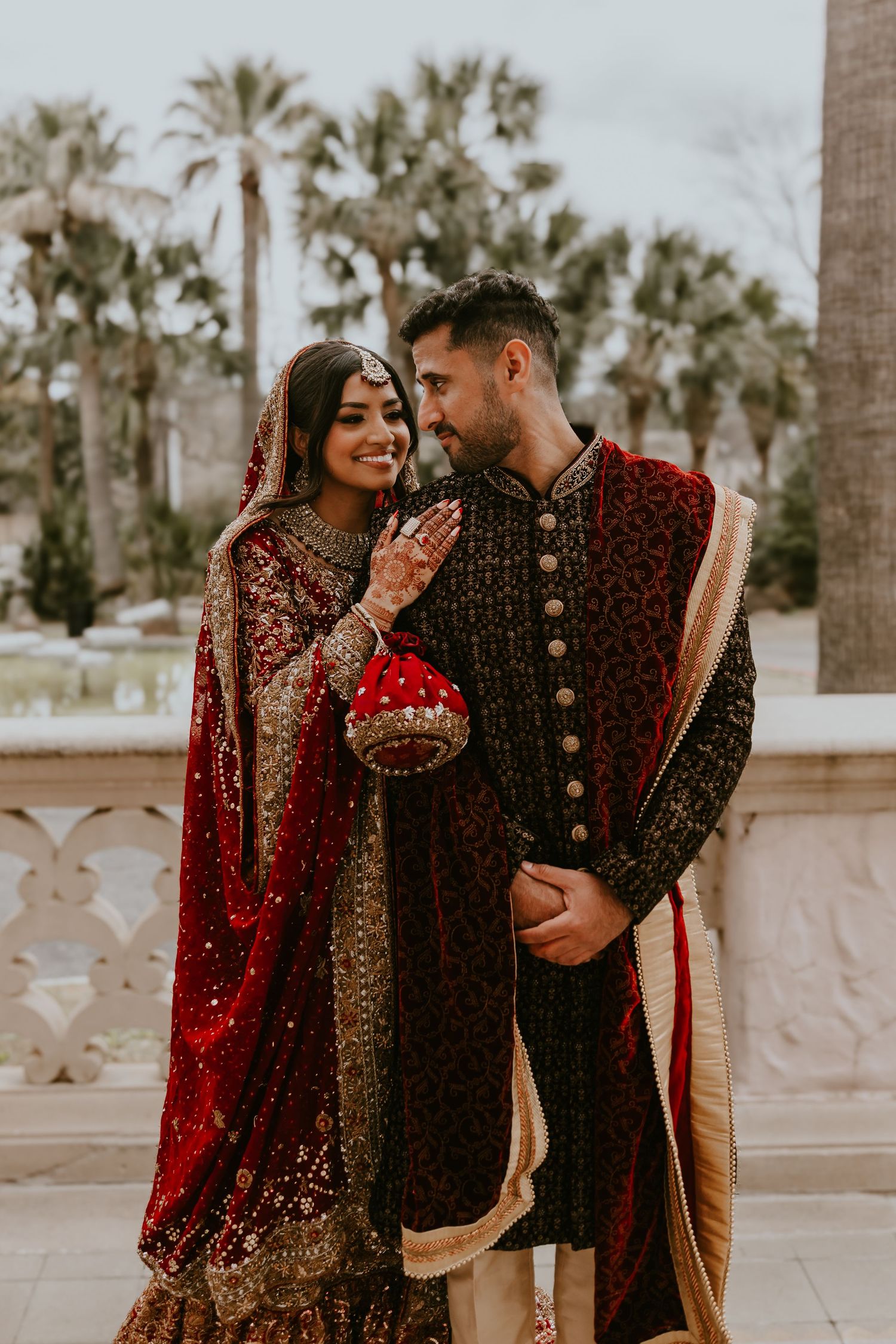 A couple wearing traditional South Asian wedding attire stands together outdoors with palm trees in the background.