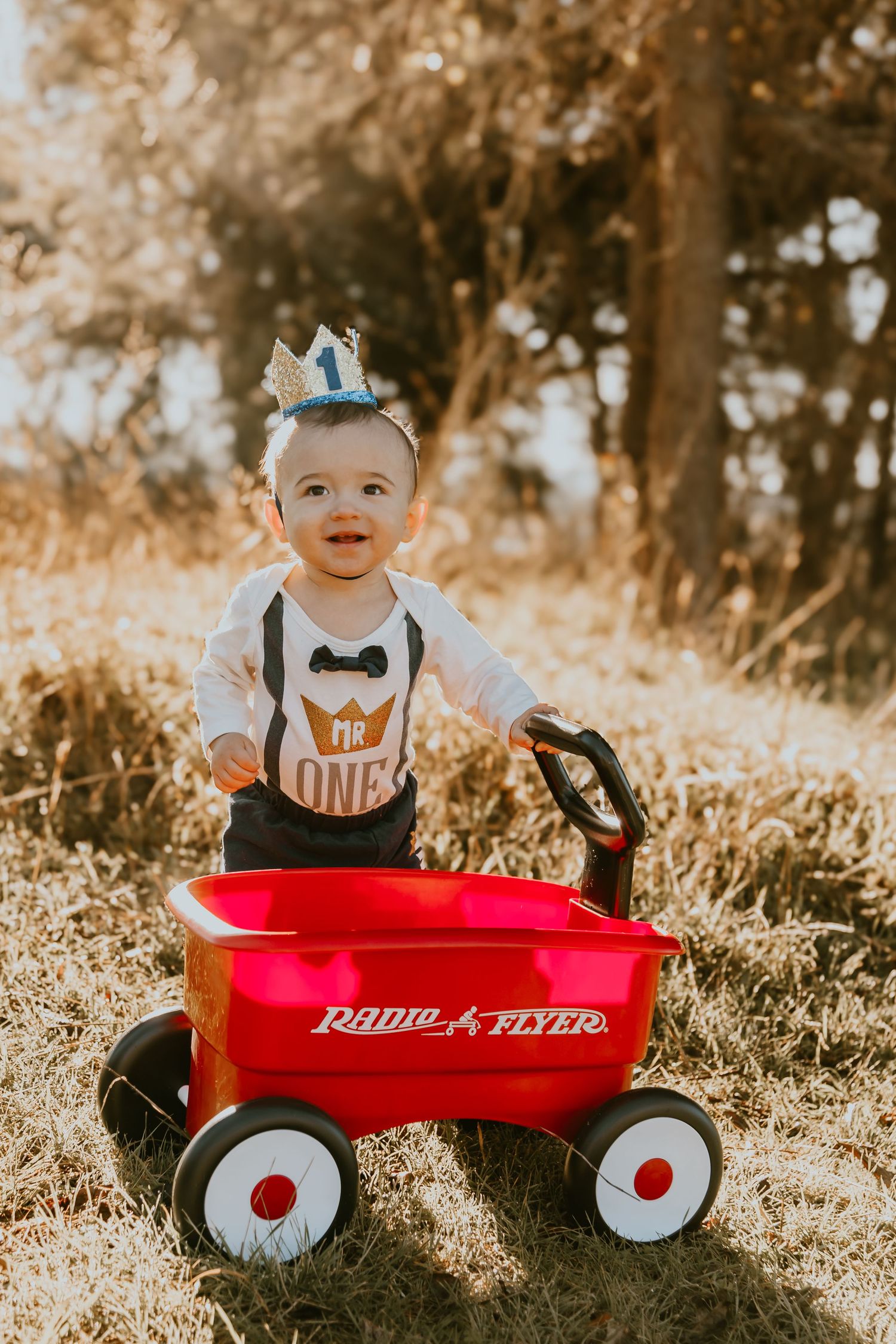 Happy toddler in suspenders and crown poses with red Radio Flyer wagon in sunlit outdoor setting.