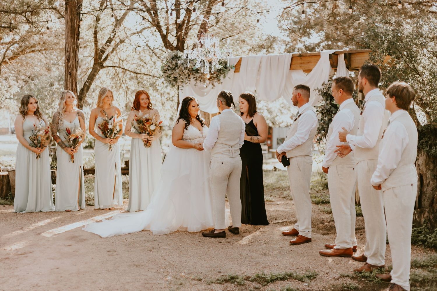 Wedding party in light blue bridesmaid dresses and white suits gathered outdoors for ceremony photos.