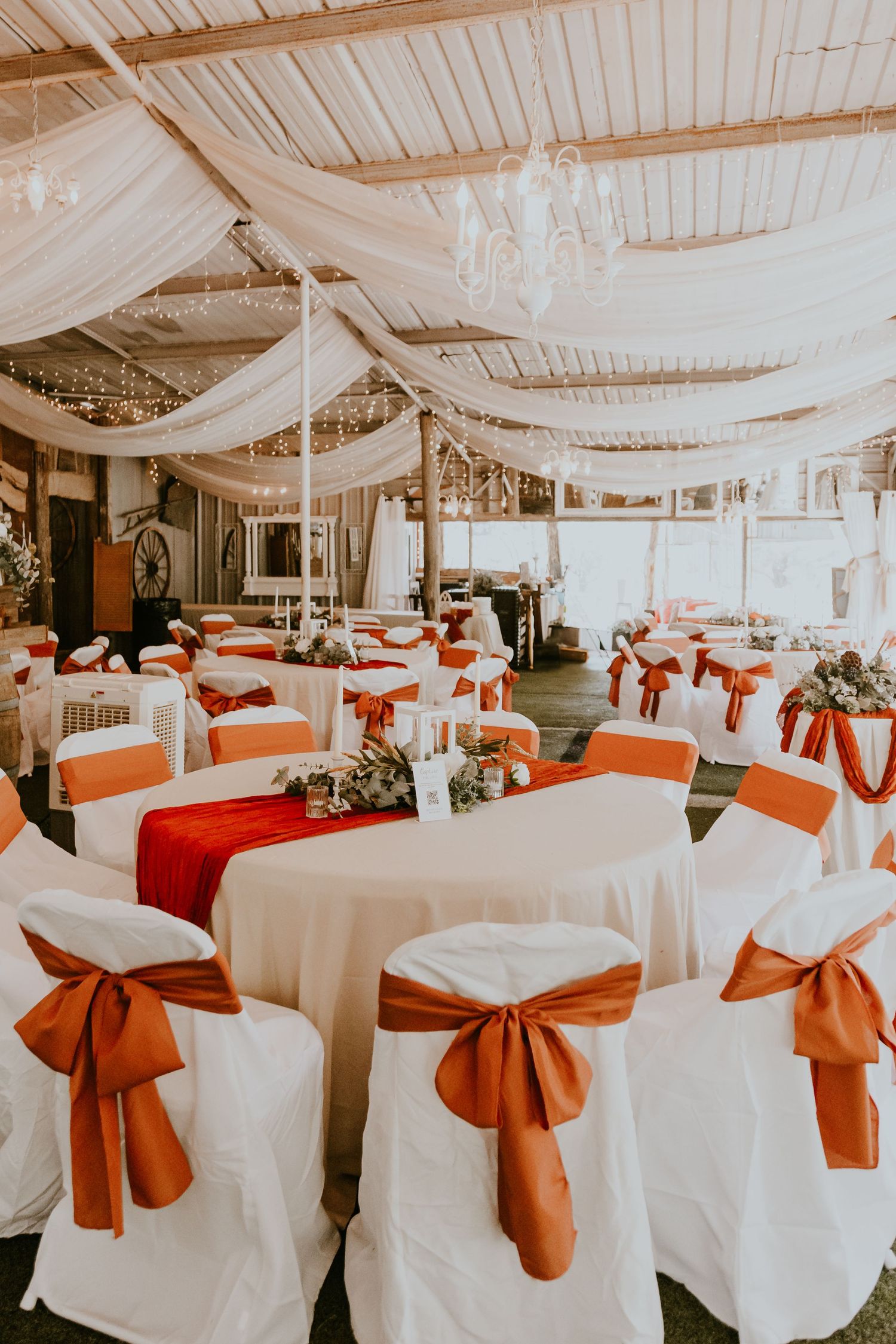 Indoor wedding reception venue decorated with white draped fabric ceiling and orange satin bows on white-covered chairs.