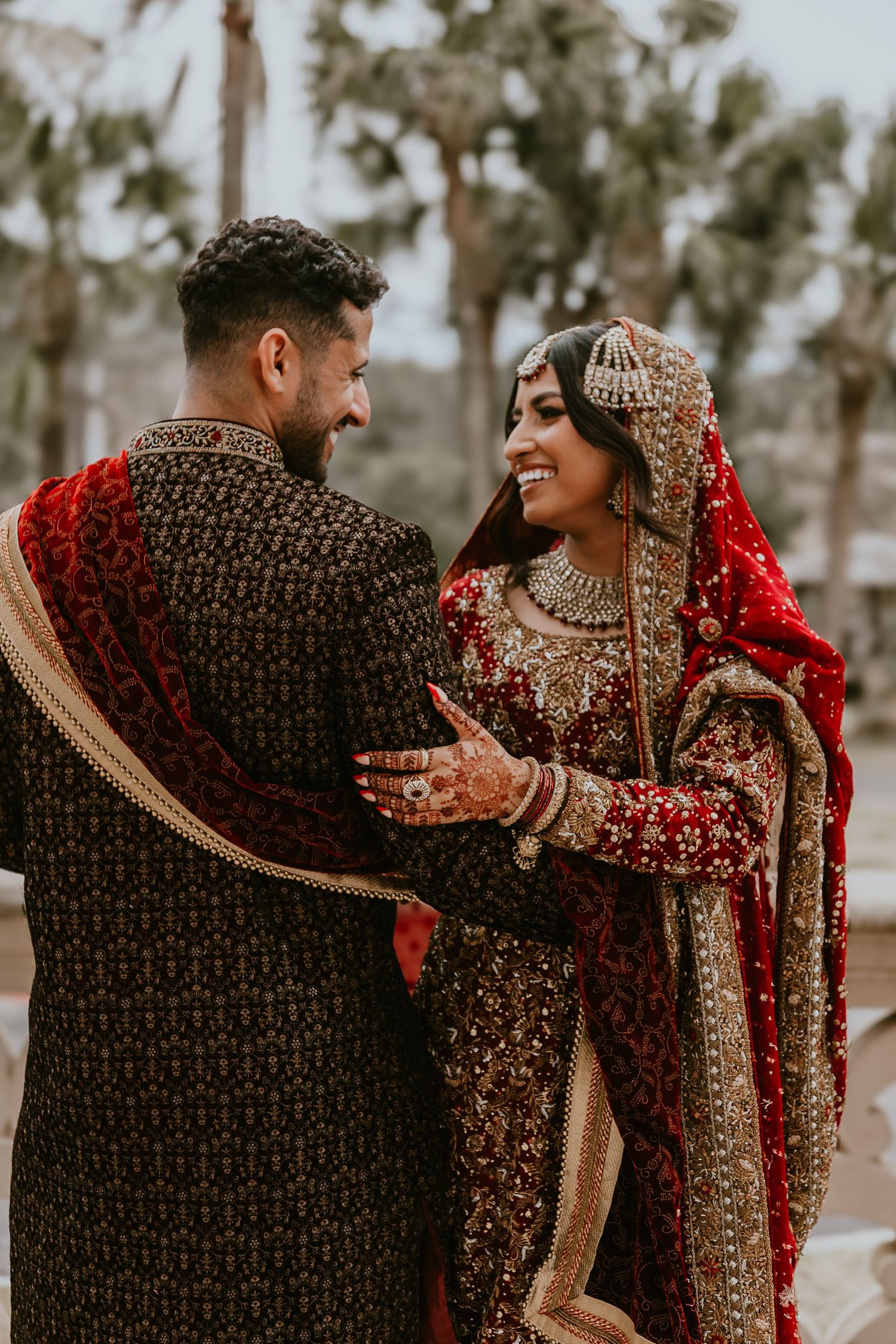 A couple in traditional South Asian wedding attire smiles at each other while wearing ornate red and gold embroidered garments.