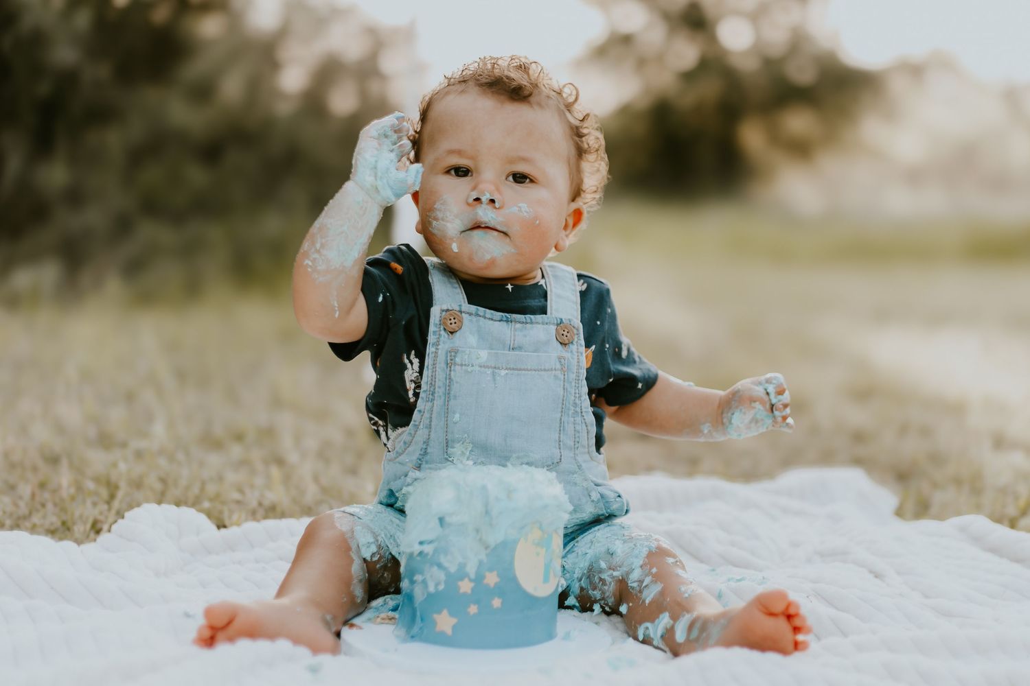 One-year-old in overalls playing with blue birthday cake during outdoor photo shoot.