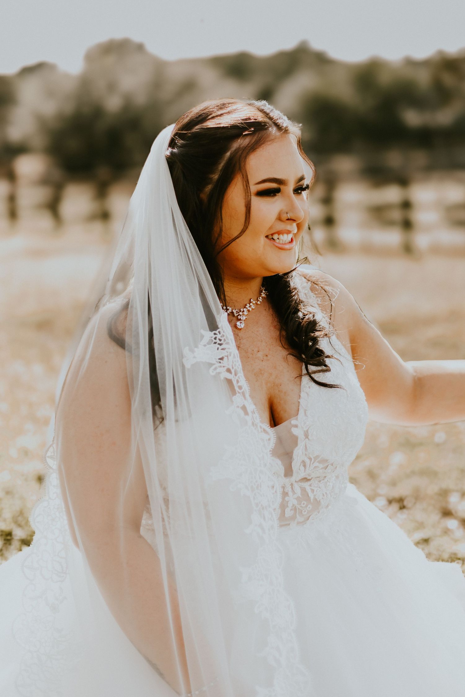 A bride wearing a lace white wedding dress and long veil smiles warmly outdoors in warm sepia-toned lighting.