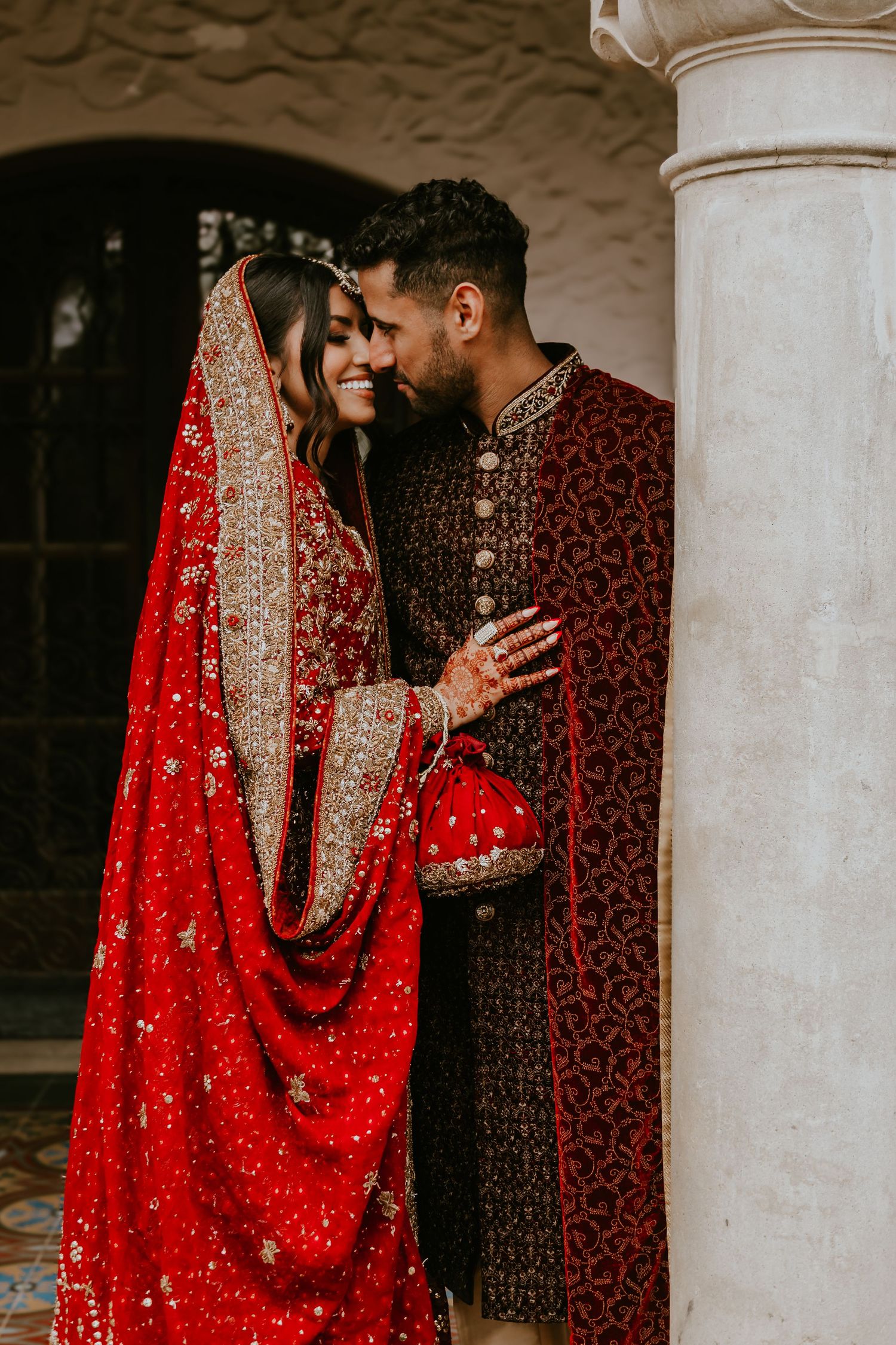 A couple in traditional South Asian wedding attire share an intimate moment by a stone pillar.