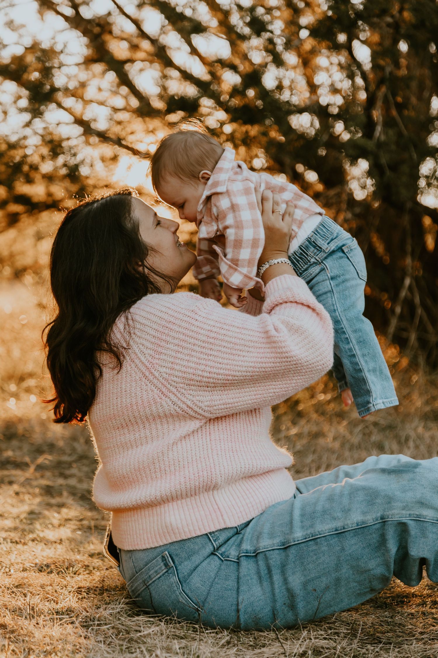 Mother in pink sweater lifts and kisses baby in casual denim during golden hour outdoor photoshoot.