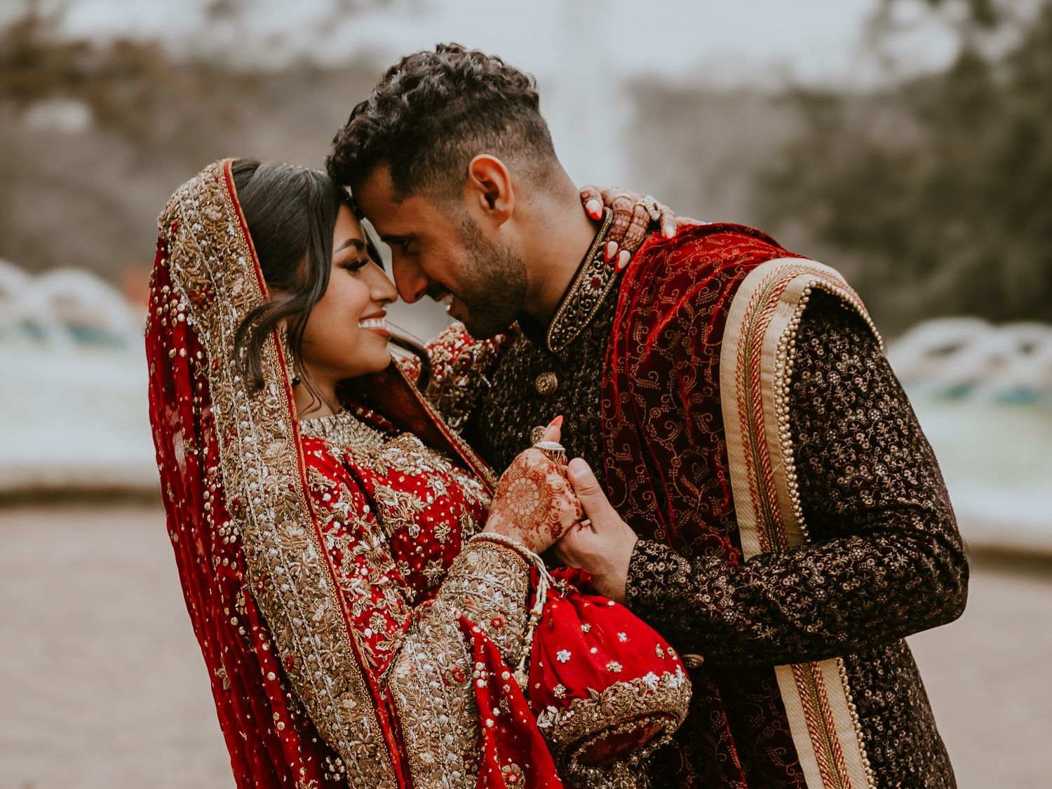 A couple in traditional South Asian wedding attire share an intimate moment in their ornate red and gold outfits.