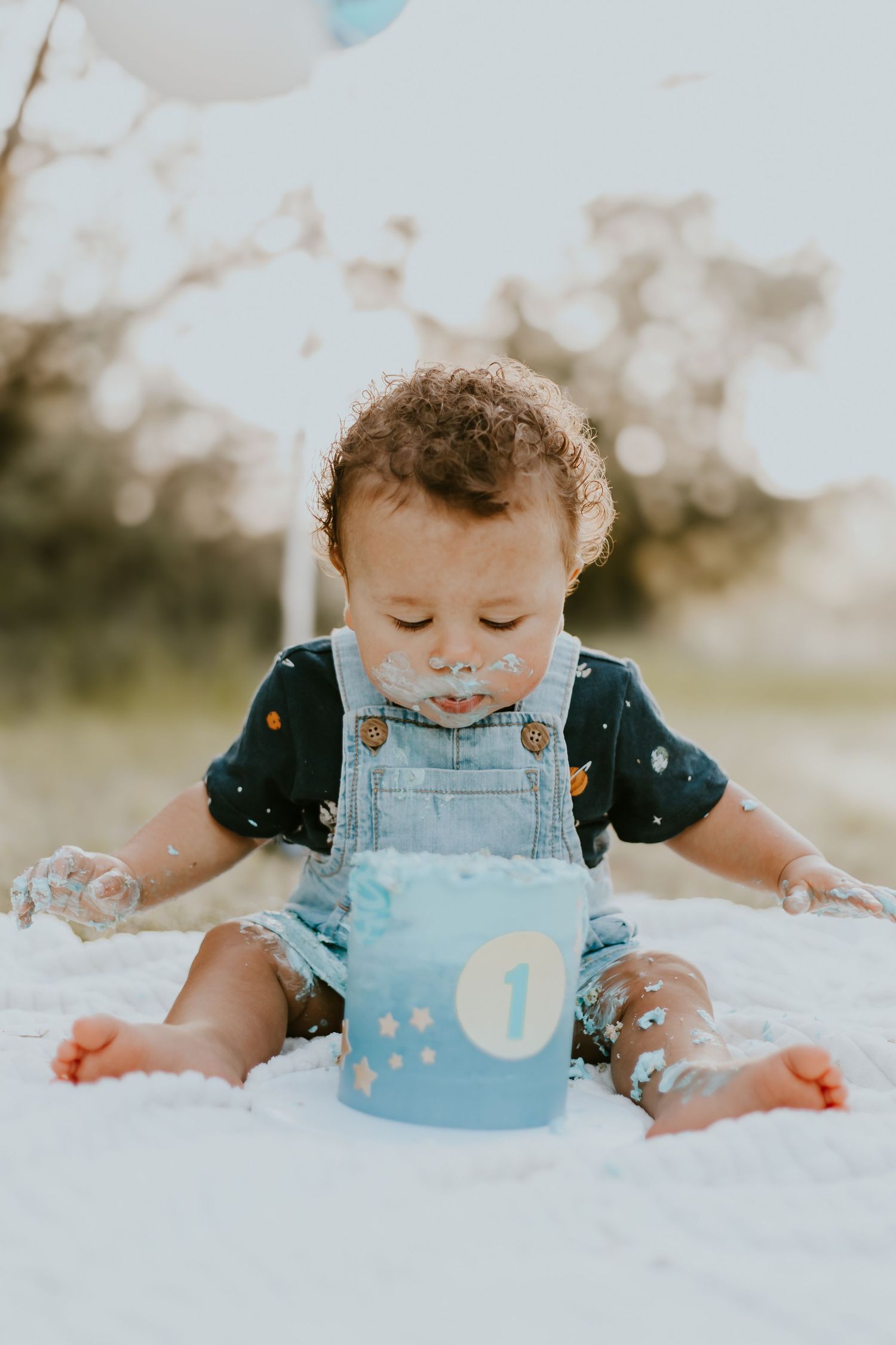 A small child in blue overalls sits on white fabric with a blue first birthday cake smeared on their face and hands.