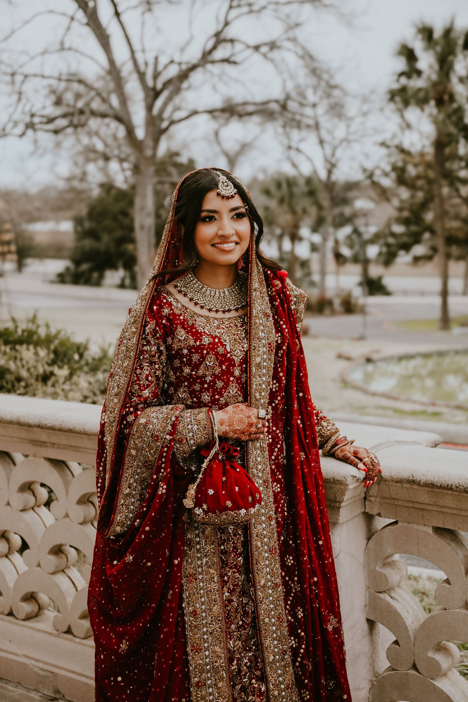 A bride in an ornate red and gold traditional South Asian lehenga poses outdoors against a scenic backdrop.
