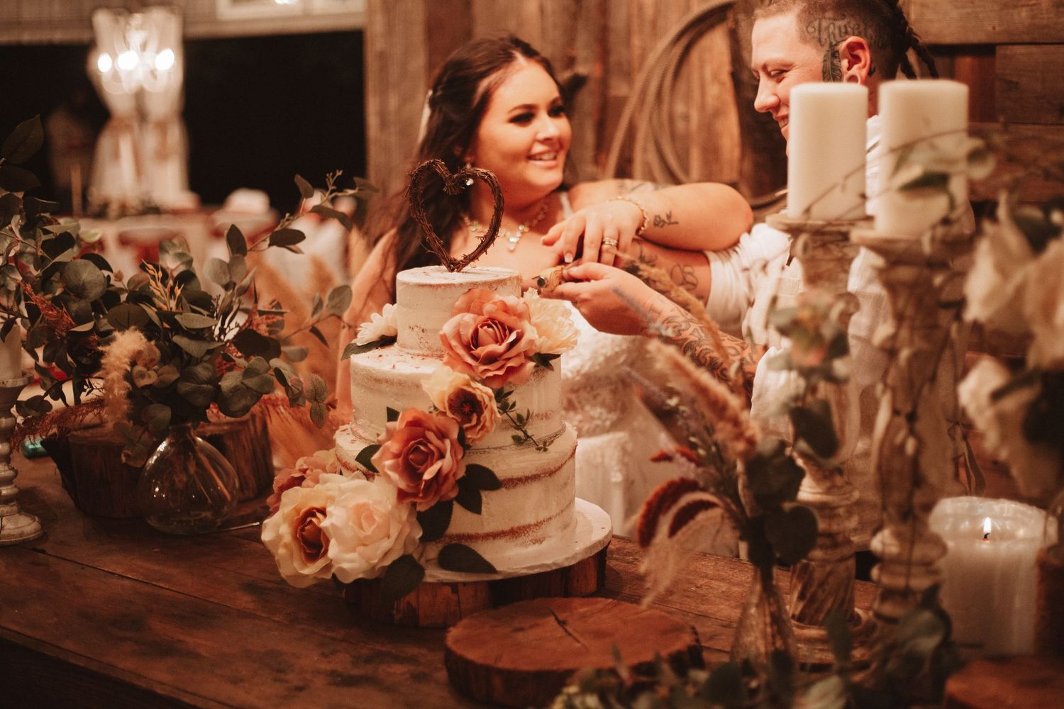 Wedding decorator arranges pink and peach roses on a rustic wooden table with glowing candles in warm lighting.