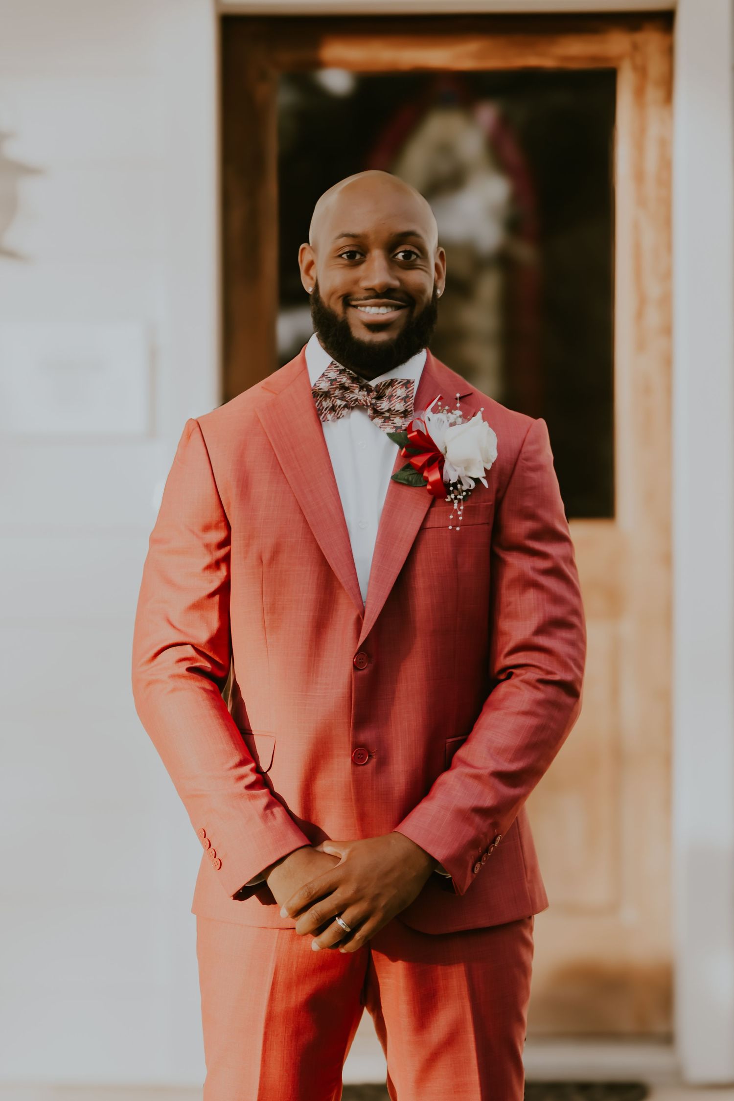 Groom in stylish coral suit with white shirt, floral bow tie and boutonniere poses against blurred indoor background.